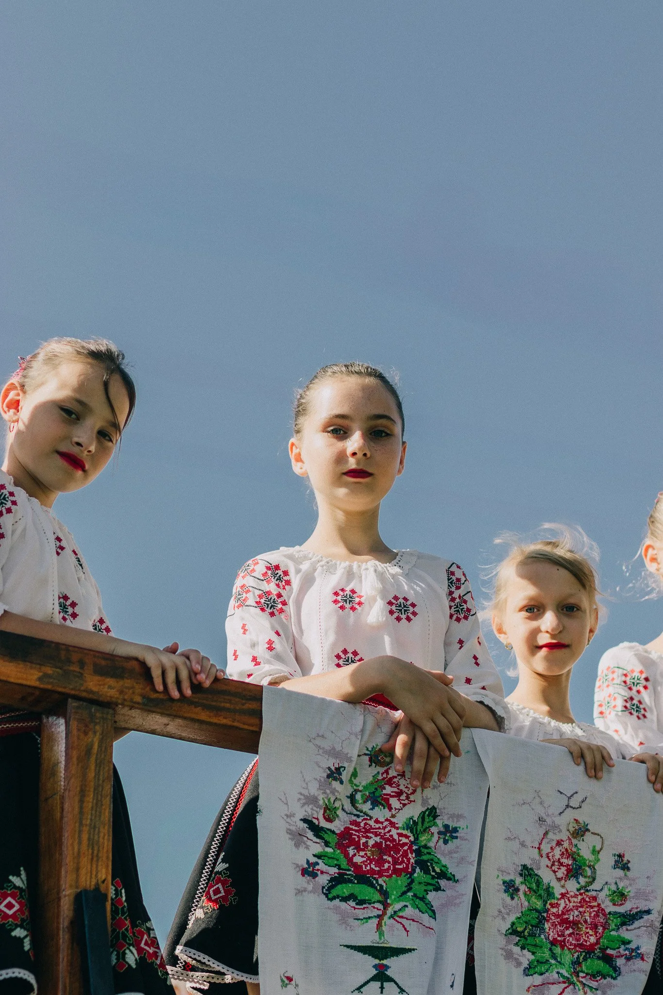 Four young girls in traditional embroidered clothing standing outdoors on a sunny day, holding embroidered cloths with floral designs.