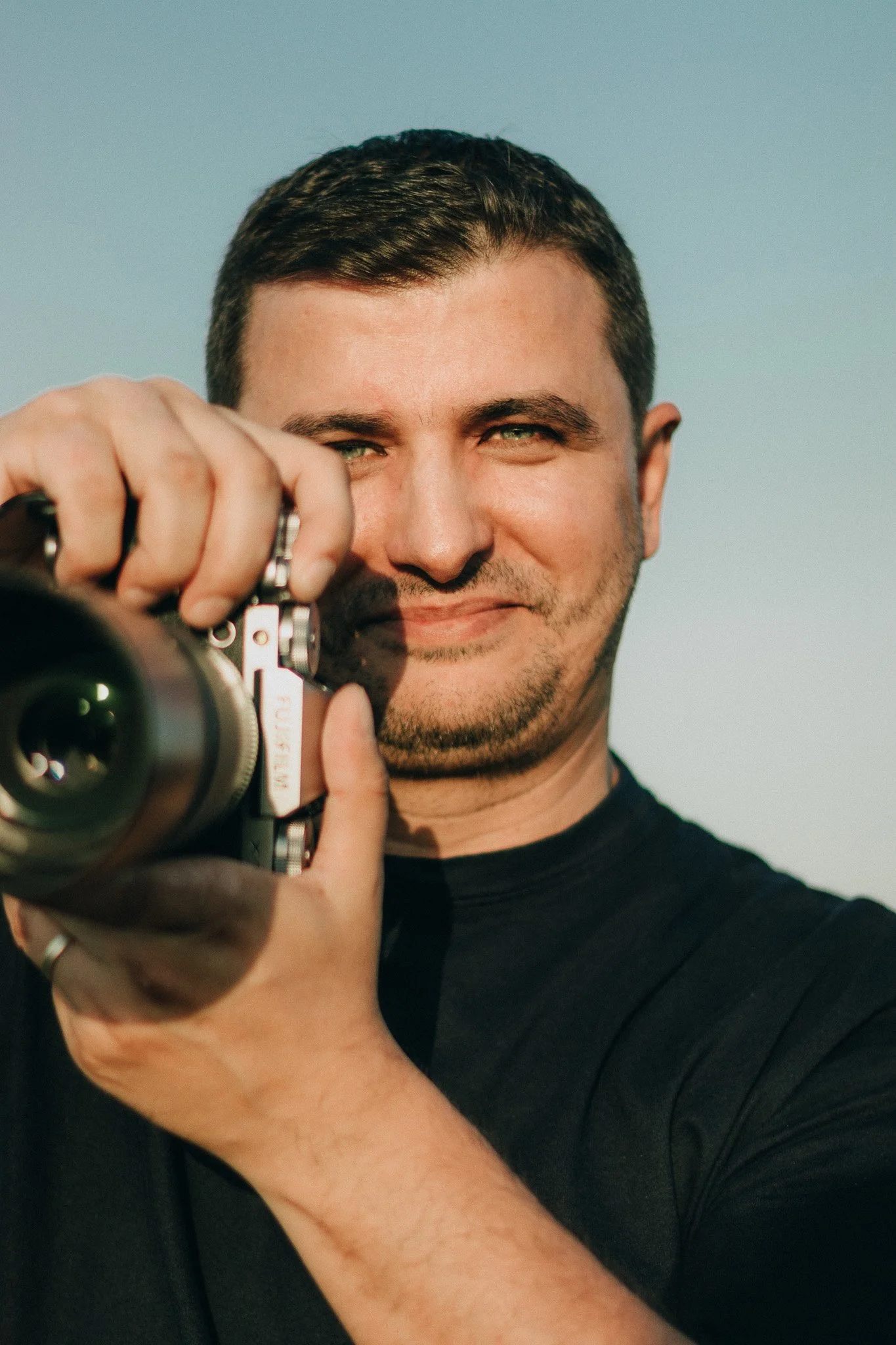 A man with short dark hair and a beard smiling while holding a camera, wearing a black shirt, with a clear sky in the background.