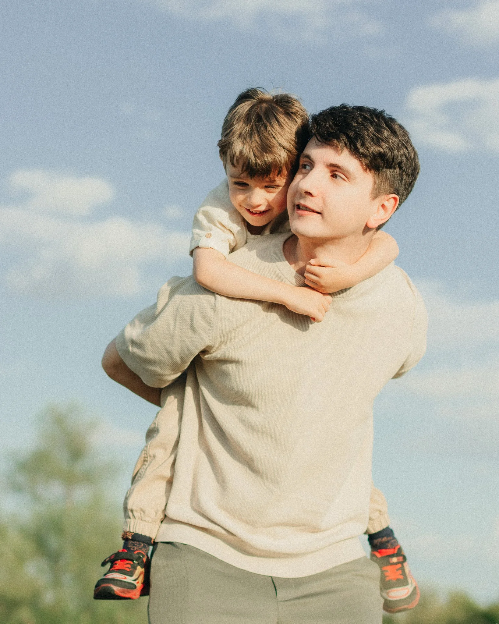A man with short dark hair giving a piggyback ride to a young boy with brown hair outdoors on a sunny day, both smiling.