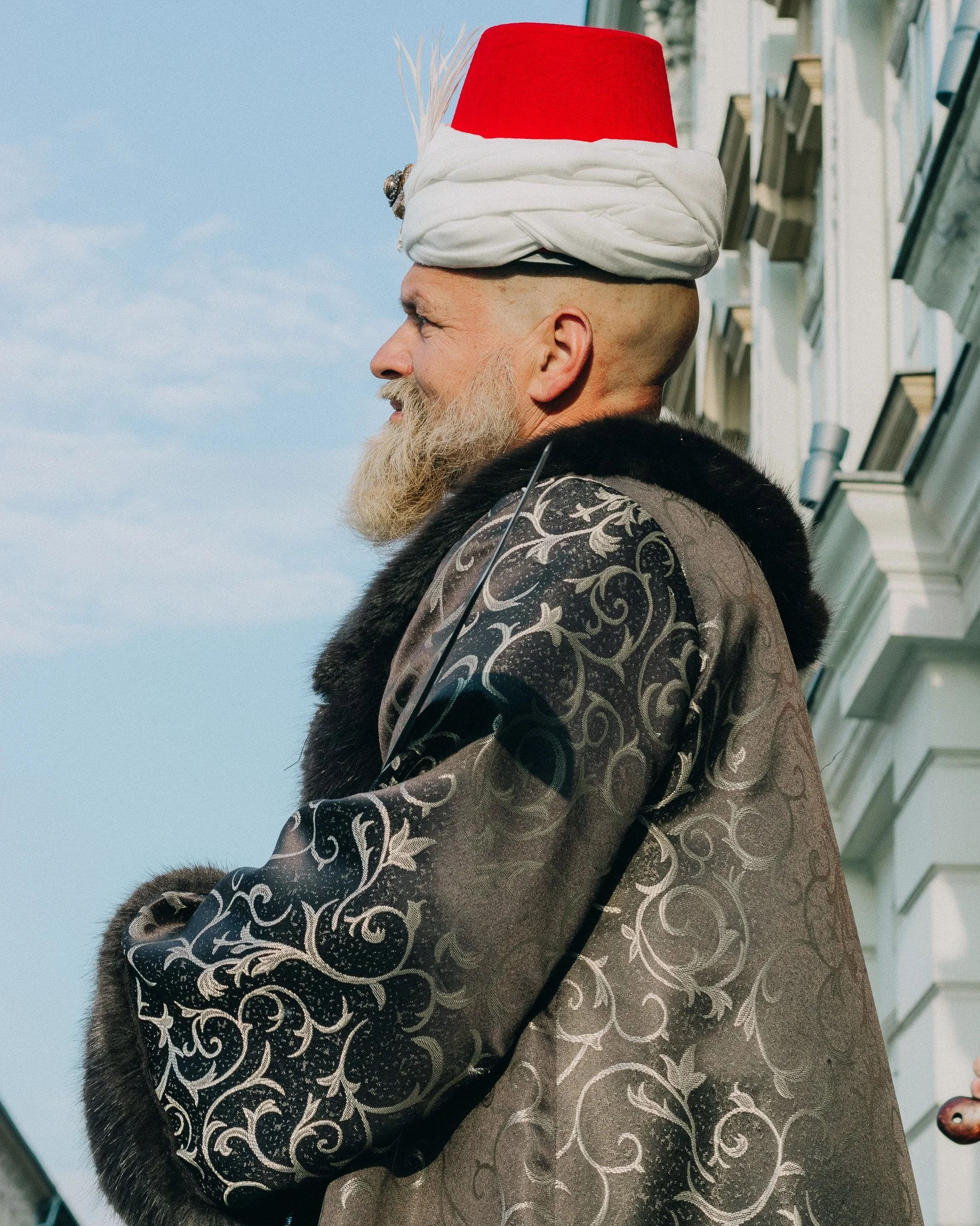 A man with a long beard wearing an ornate black and silver patterned coat, a white turban with a red top, and a black fur collar, standing outdoors against a blue sky and white building.