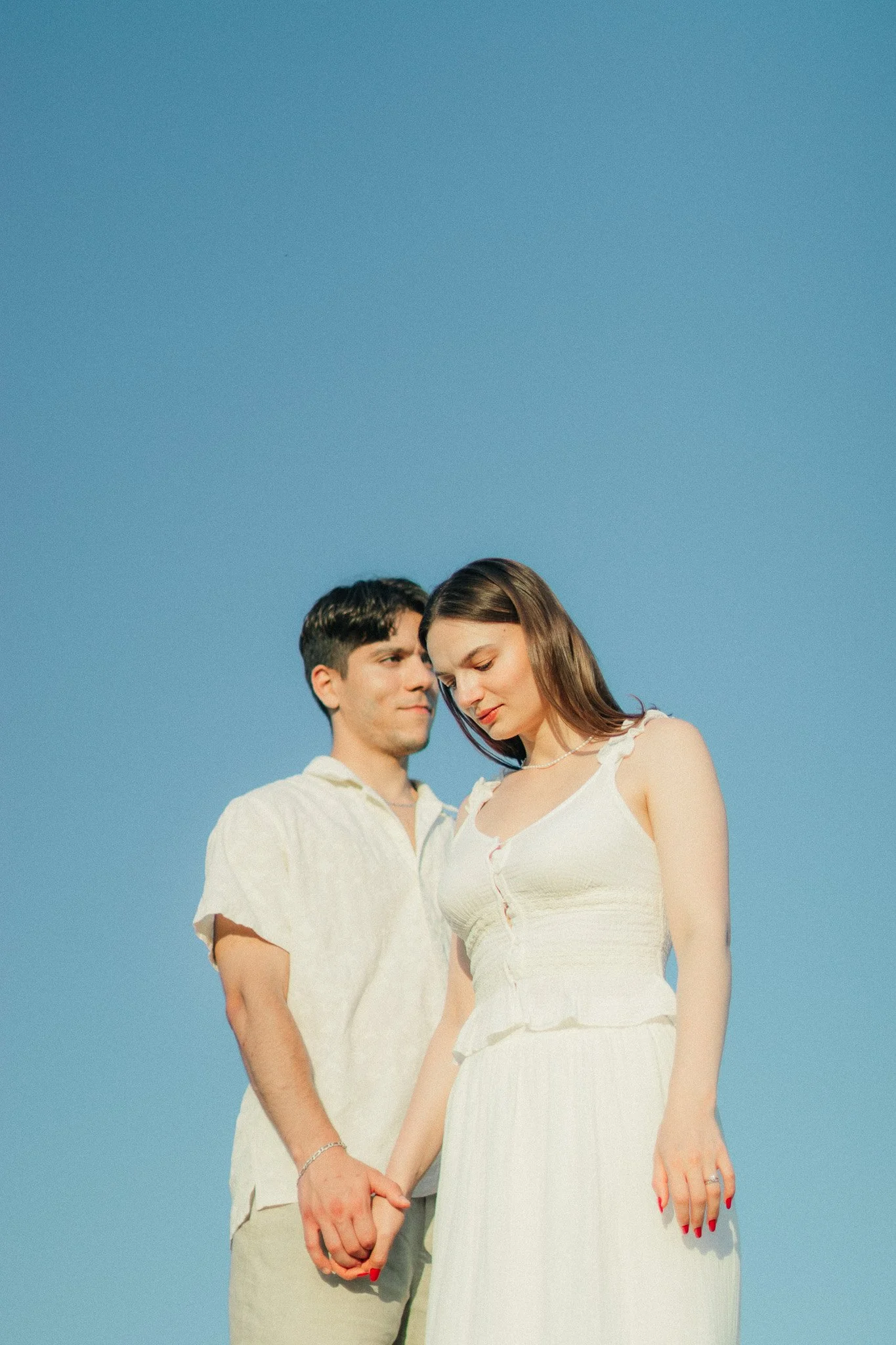 A young couple holding hands, standing outdoors under a clear blue sky, with the woman looking down softly and the man smiling gently.