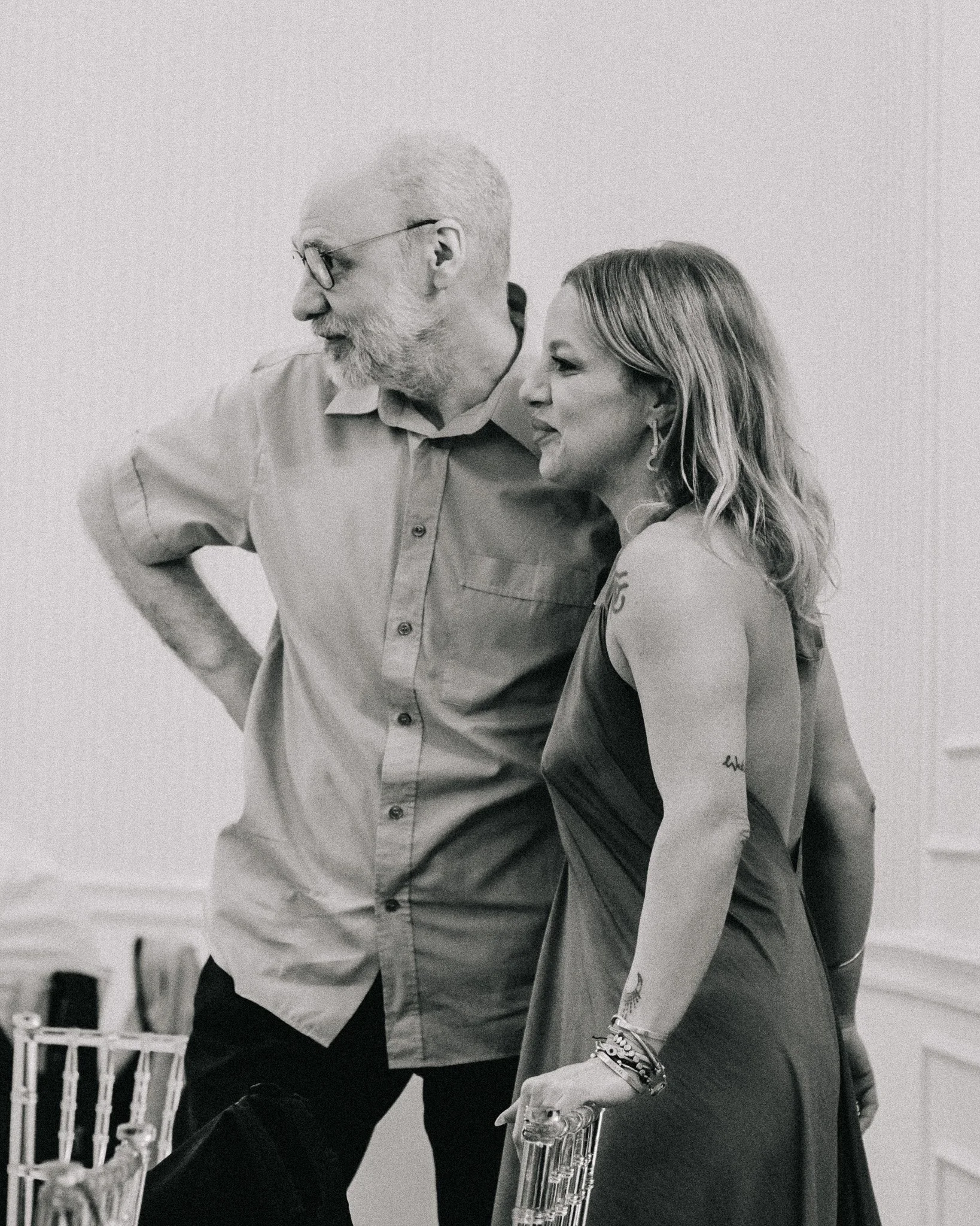 A black and white photograph of an older man with glasses and a beard standing next to a woman with wavy hair. They are indoors, likely at a social event, and appear to be engaged in conversation or listening to something.