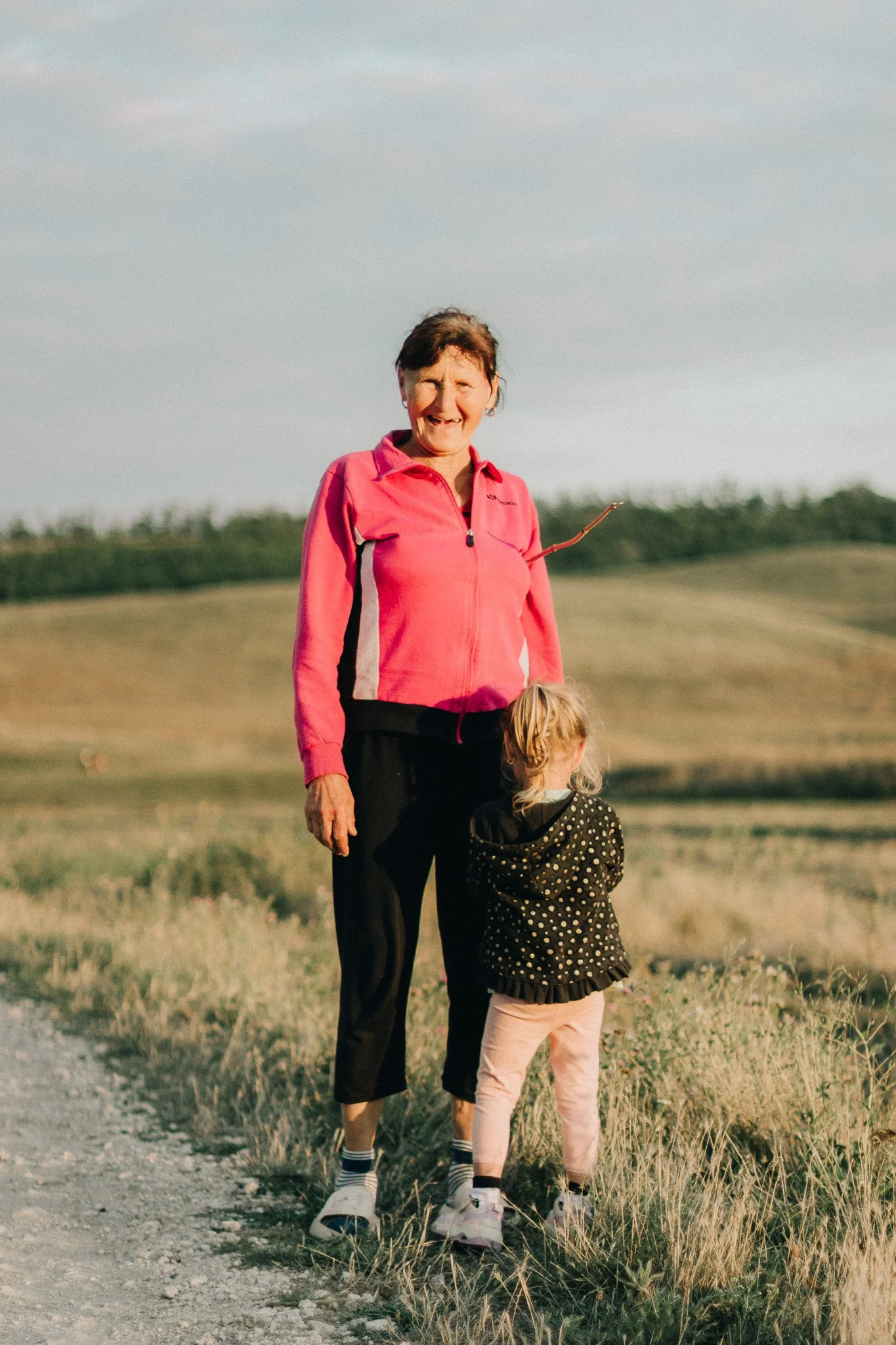An elderly woman wearing a pink jacket and black pants walking outdoors on a trail with a young girl in a black polka-dot jacket and pink pants, both facing away from the camera, in a grassy, hilly landscape.