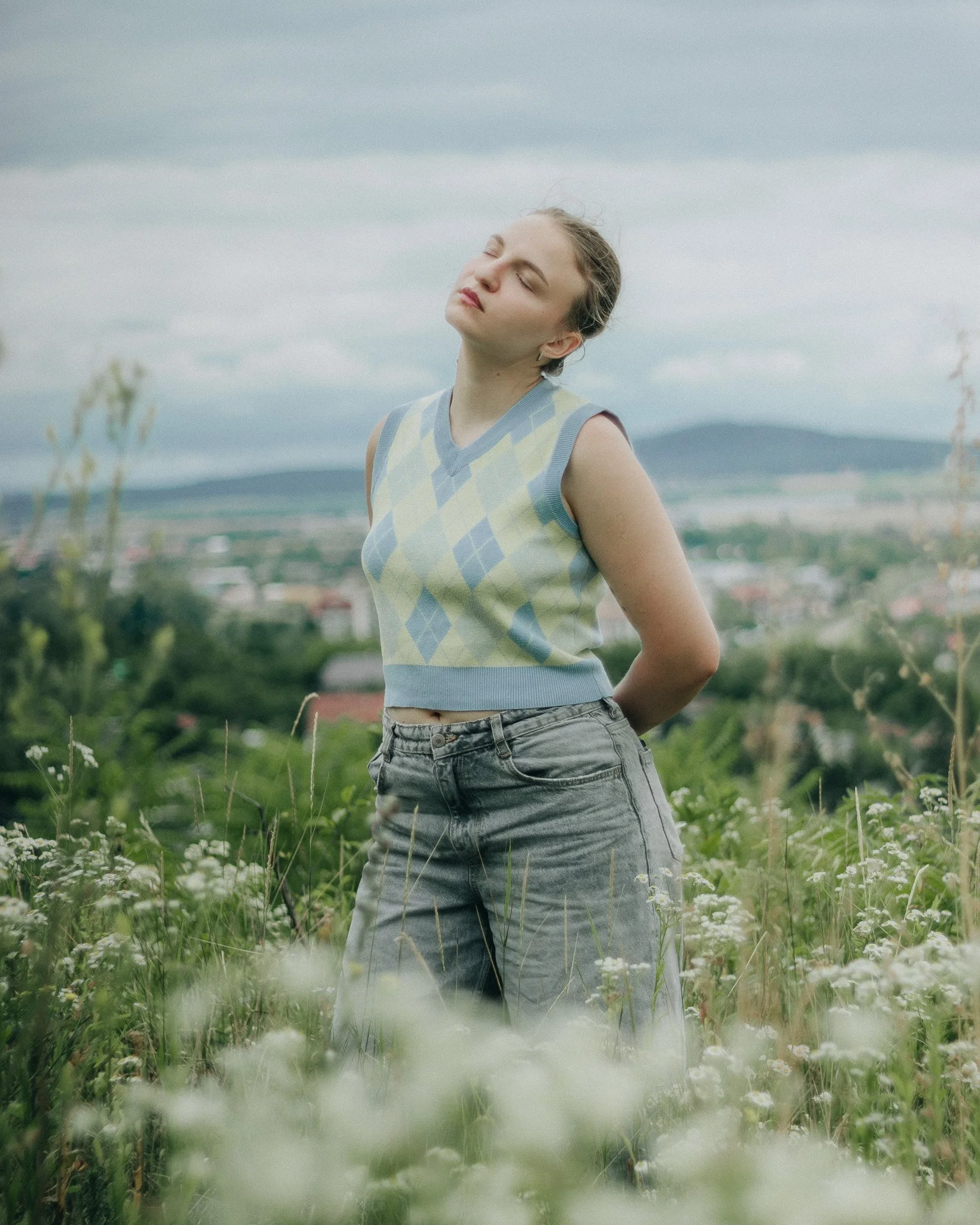 A woman in a sleeveless sweater and jeans standing in a field of white flowers with closed eyes and a peaceful expression, with a cloudy sky and distant landscape in the background.