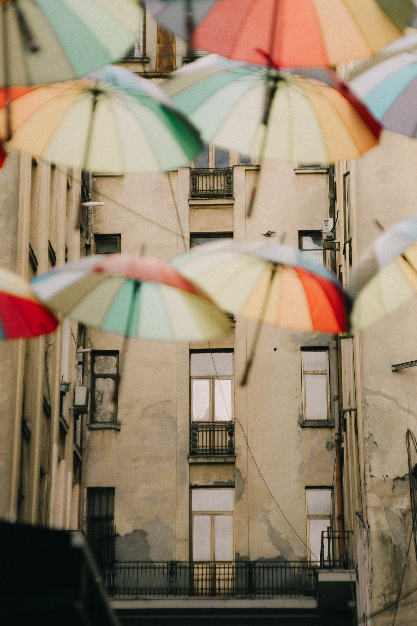Colorful umbrellas hanging above a worn, beige-colored building with multiple windows and small balconies.