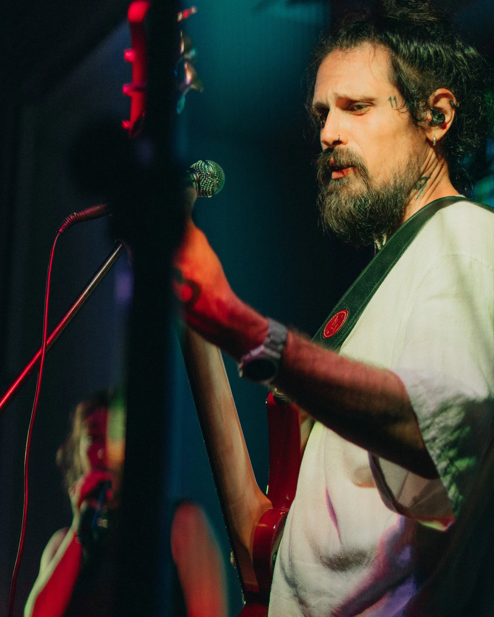 A man with a beard and tattoos, wearing a white T-shirt, plays an acoustic guitar on stage with a microphone in front of him.