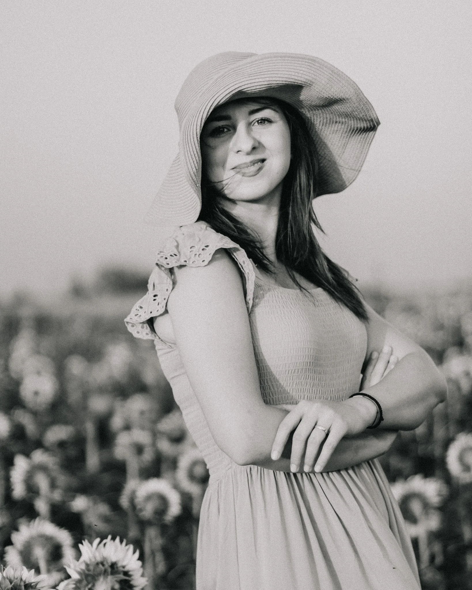 A woman wearing a large sunhat and a dress with lace details on the shoulders, standing in a field of sunflowers, smiling and crossing her arms.