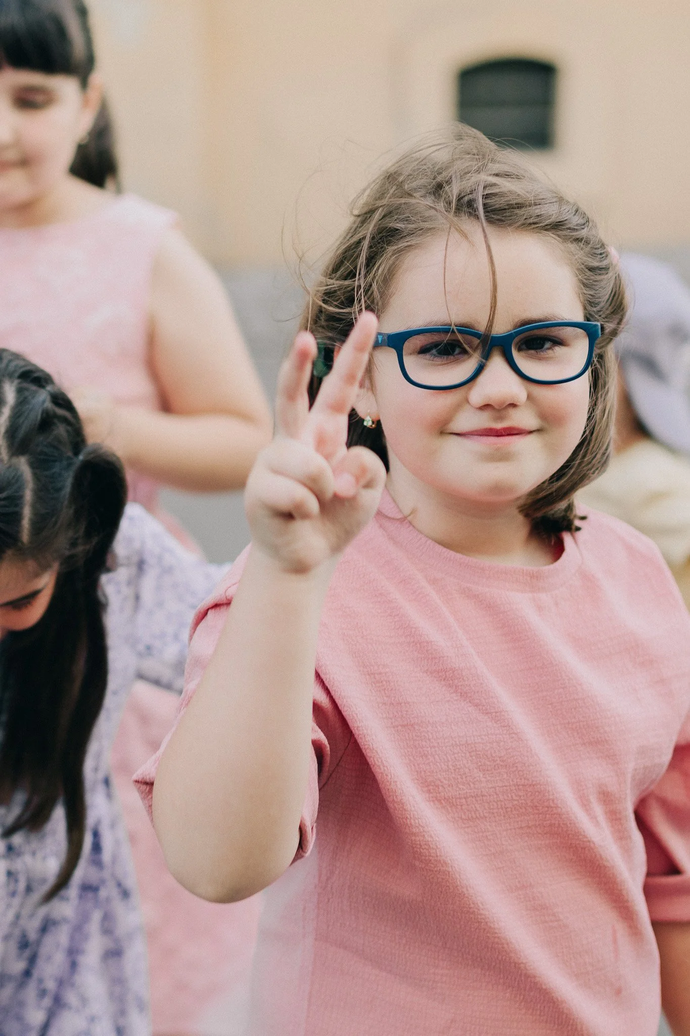 A girl with glasses and a pink shirt making a peace sign and smiling at the camera.