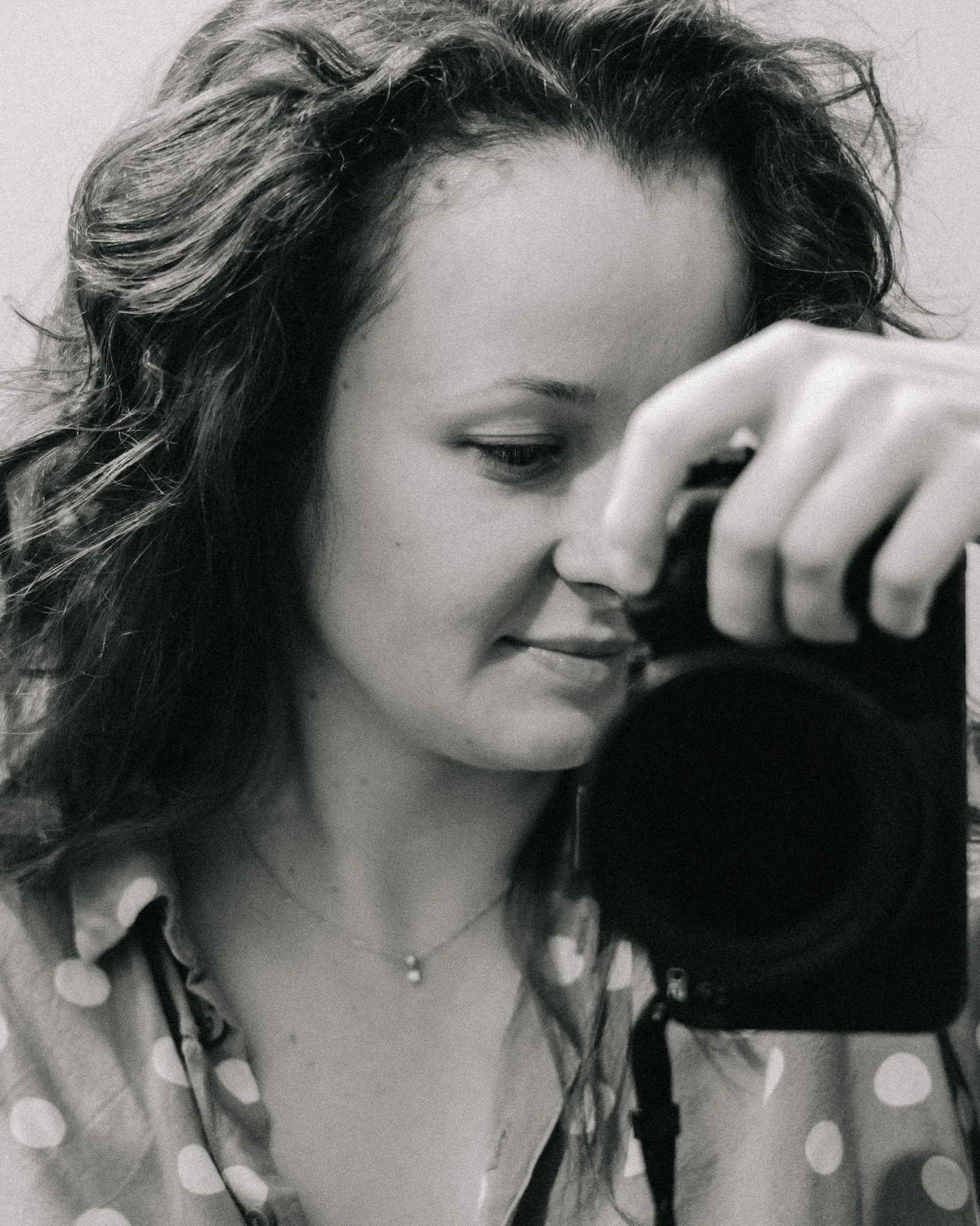 A young woman with wavy hair taking a selfie with a camera, wearing a polka dot shirt and a necklace, in black and white.