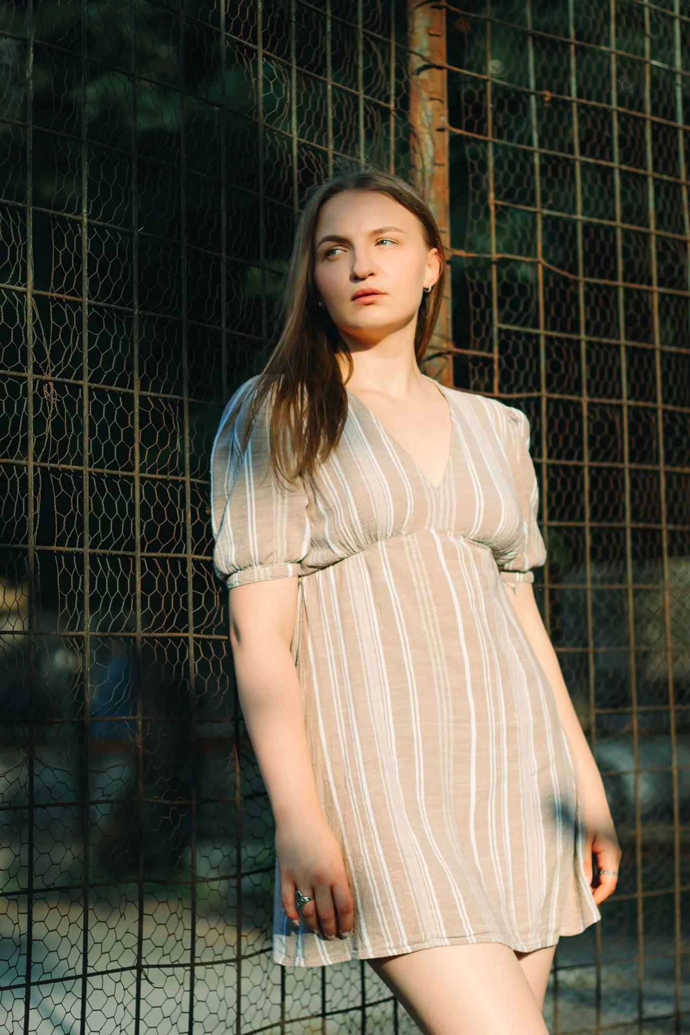 Young woman standing next to a chain-link fence, gazing to the side, wearing a beige striped dress during daylight.