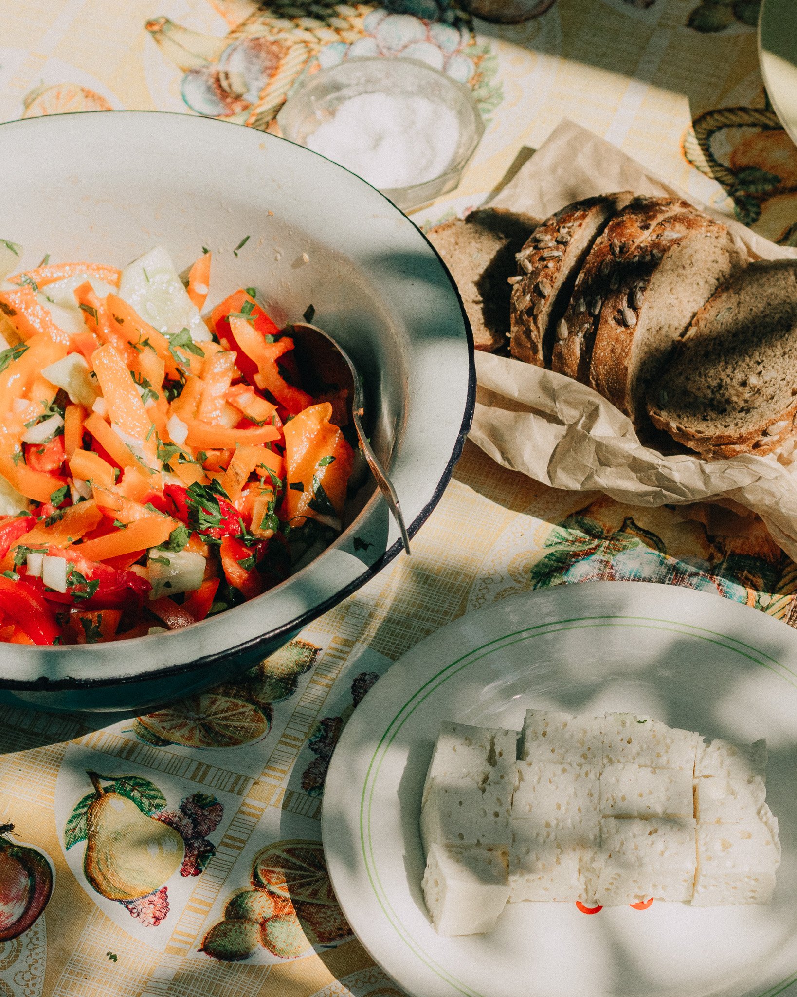 A table with a large salad bowl containing chopped tomatoes, cucumbers, and herbs, a plate of sliced cheese or bread, a basket of sliced multigrain bread, and a small bowl of salt.