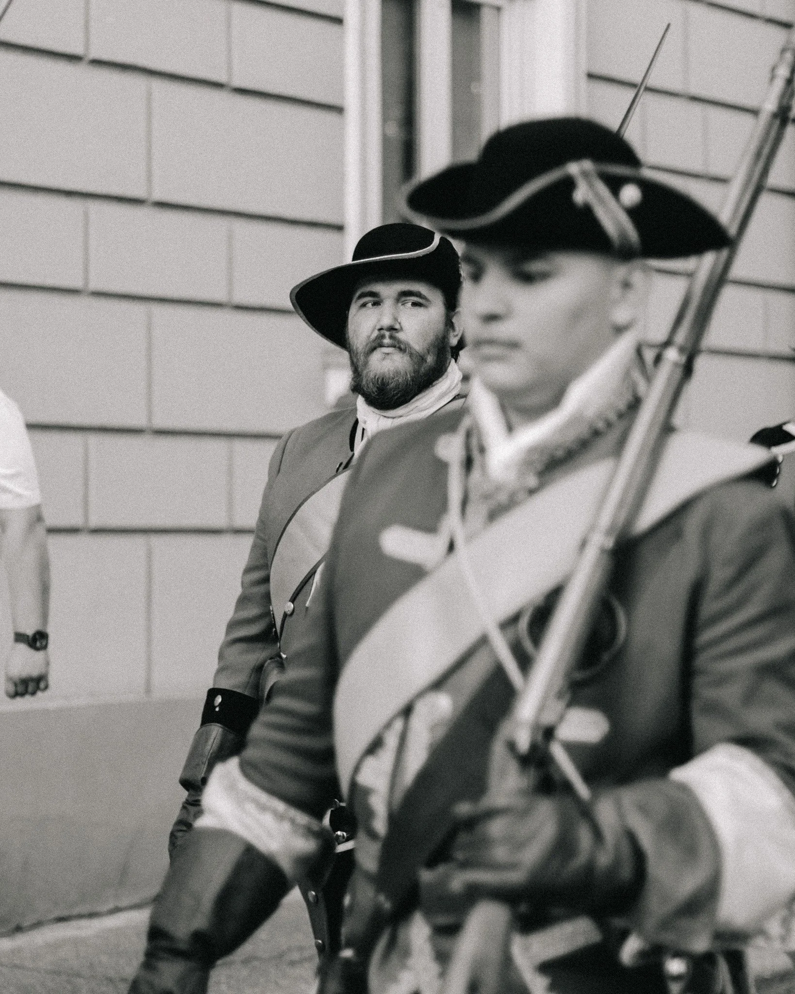Two men dressed as revolutionary soldiers from the American Revolutionary War, wearing tricorn hats and period clothing, one with a musket, sitting outdoors with a brick building in the background.