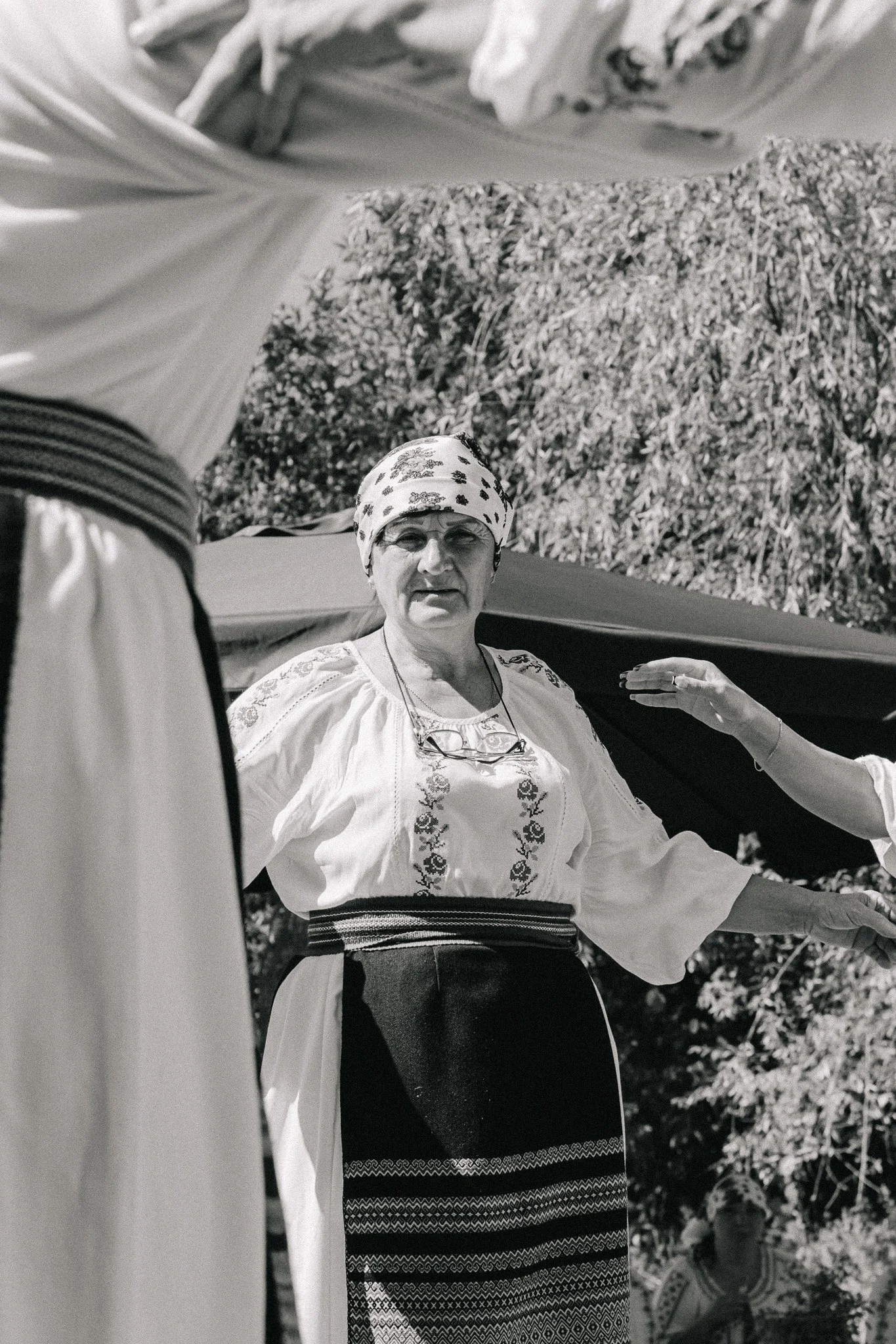 A woman in traditional clothing standing outdoors, with another person's arm partially visible, and trees in the background.