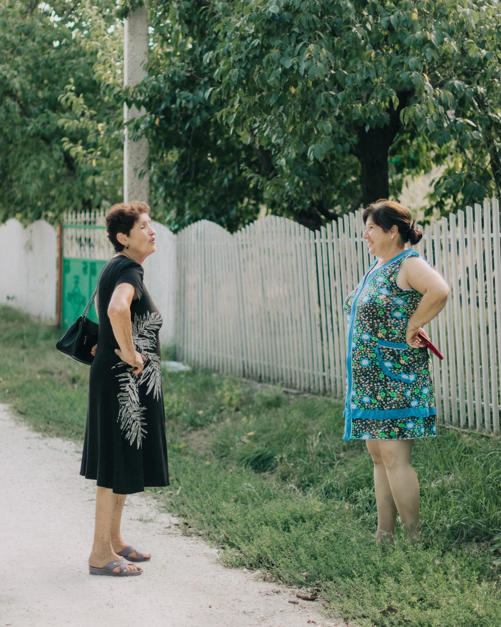 Two women standing outdoors on a grassy area beside a white picket fence, engaged in conversation, with trees in the background.
