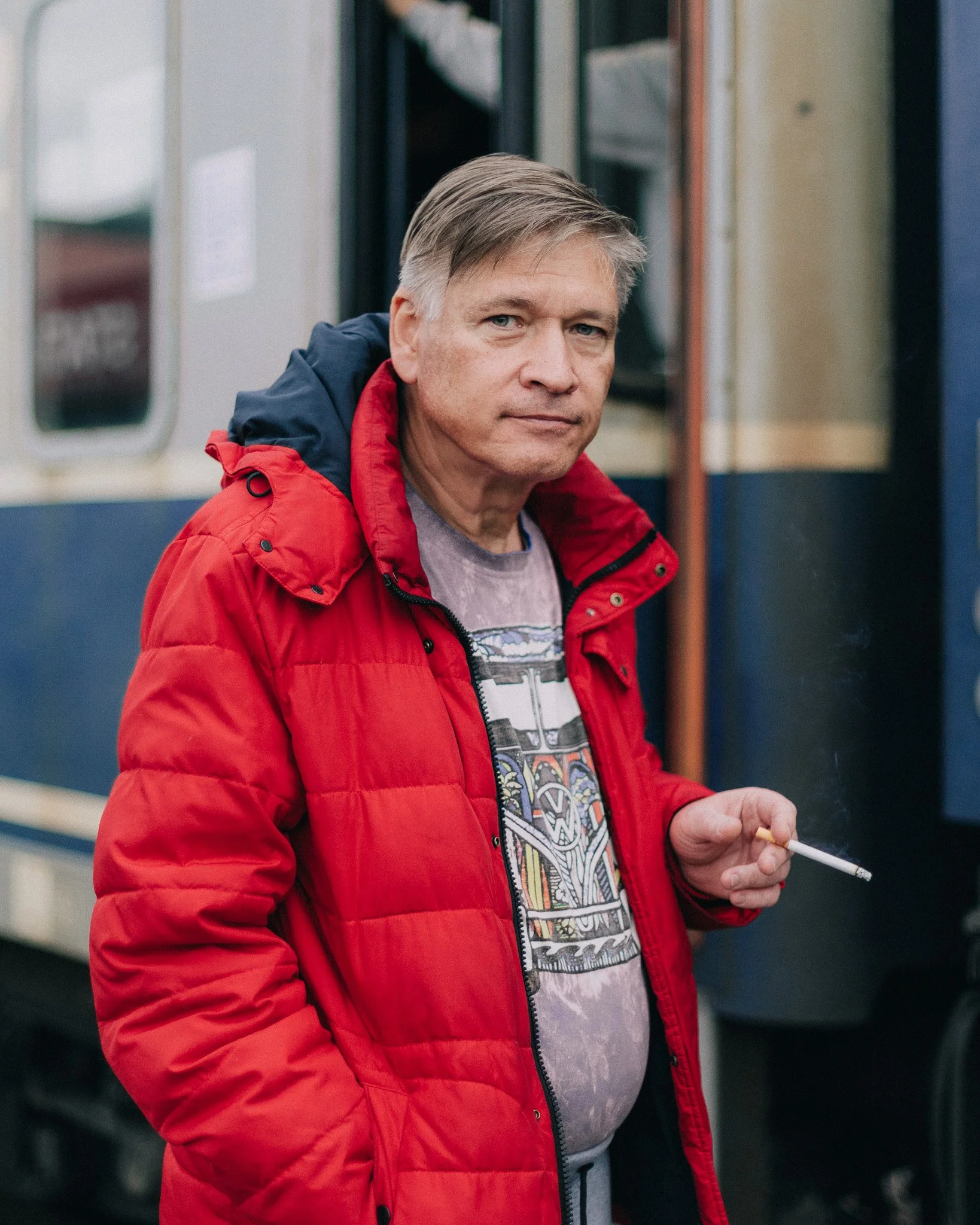 A man with gray hair wearing a red jacket holding a cigarette, standing near a train.