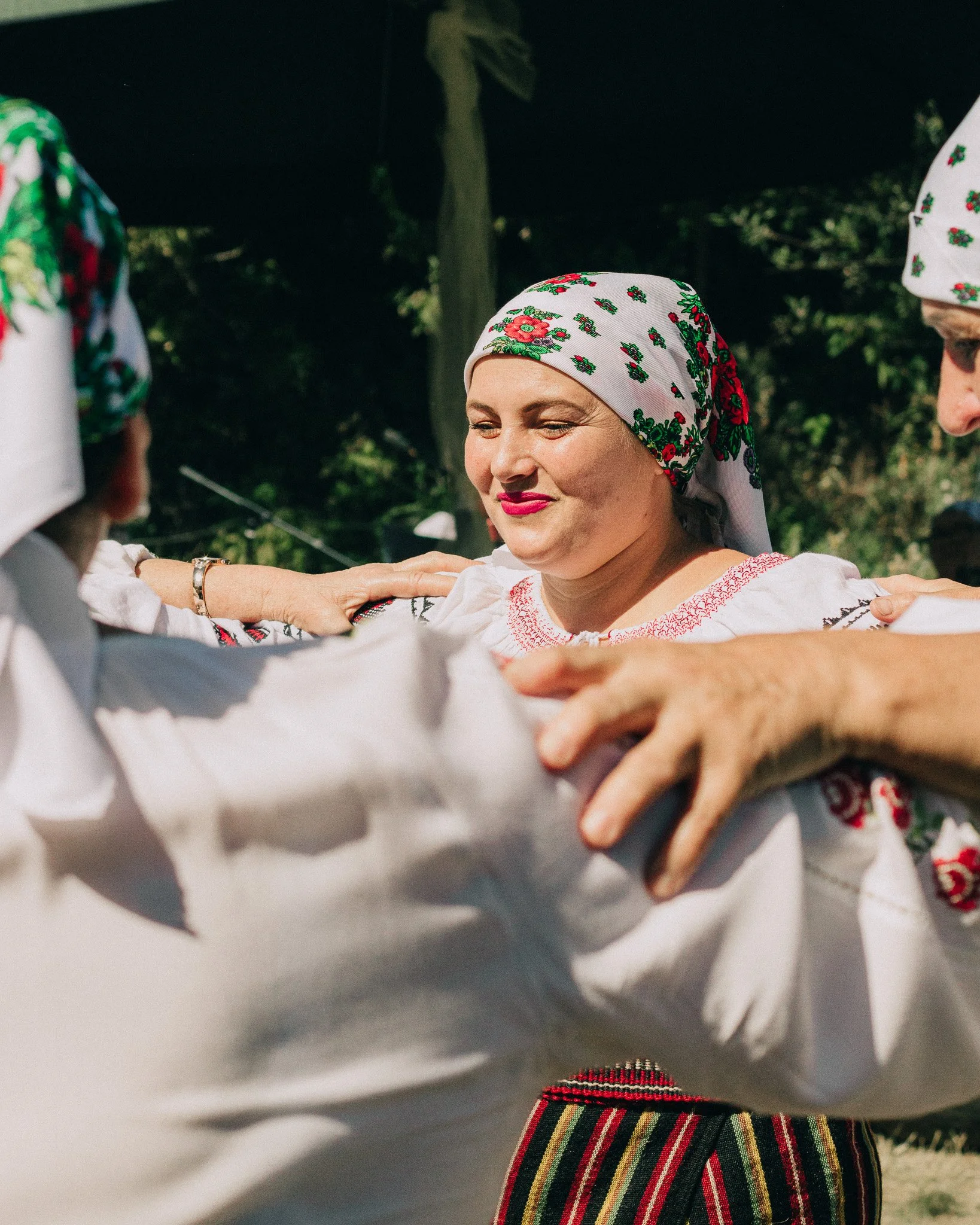Women dressed in traditional folk costumes, dancing outdoors, with one woman in the center wearing a headscarf with floral patterns, a white embroidered blouse, and a striped skirt.
