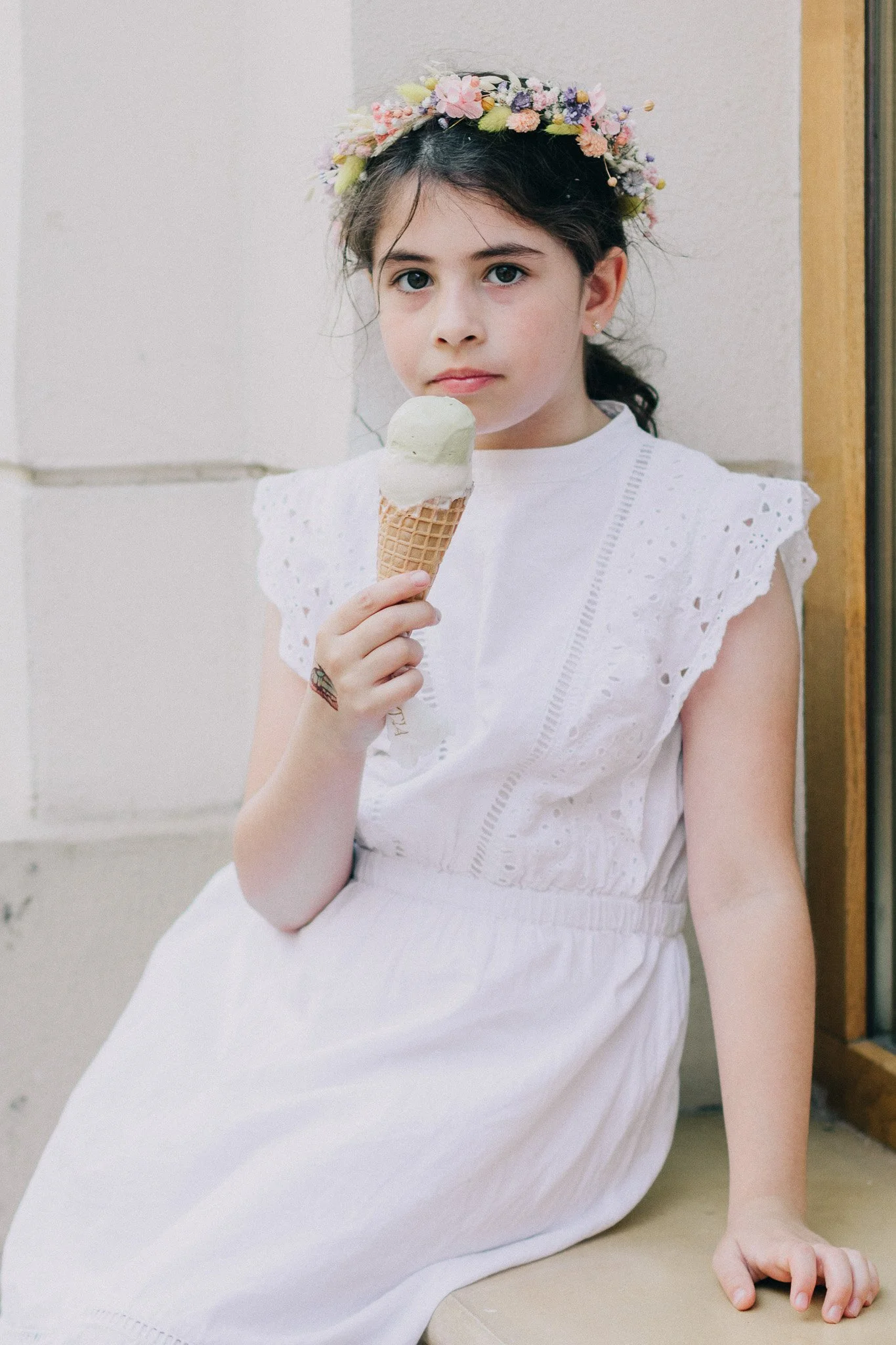 A young girl with a floral headband, wearing a white dress, is sitting against a wall and holding an ice cream cone.