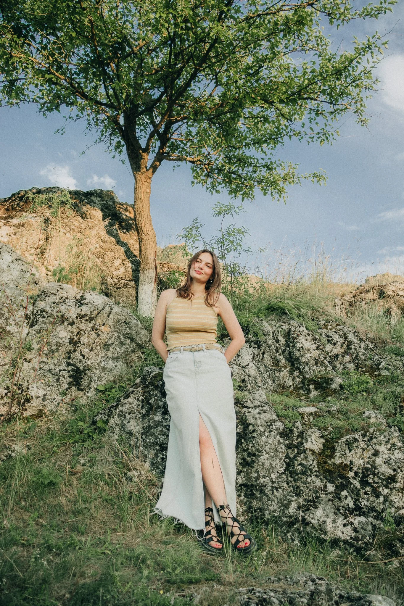 A young woman standing on rocks outdoors under a large tree, wearing a beige striped tank top and a light-colored skirt with a slit, smiling