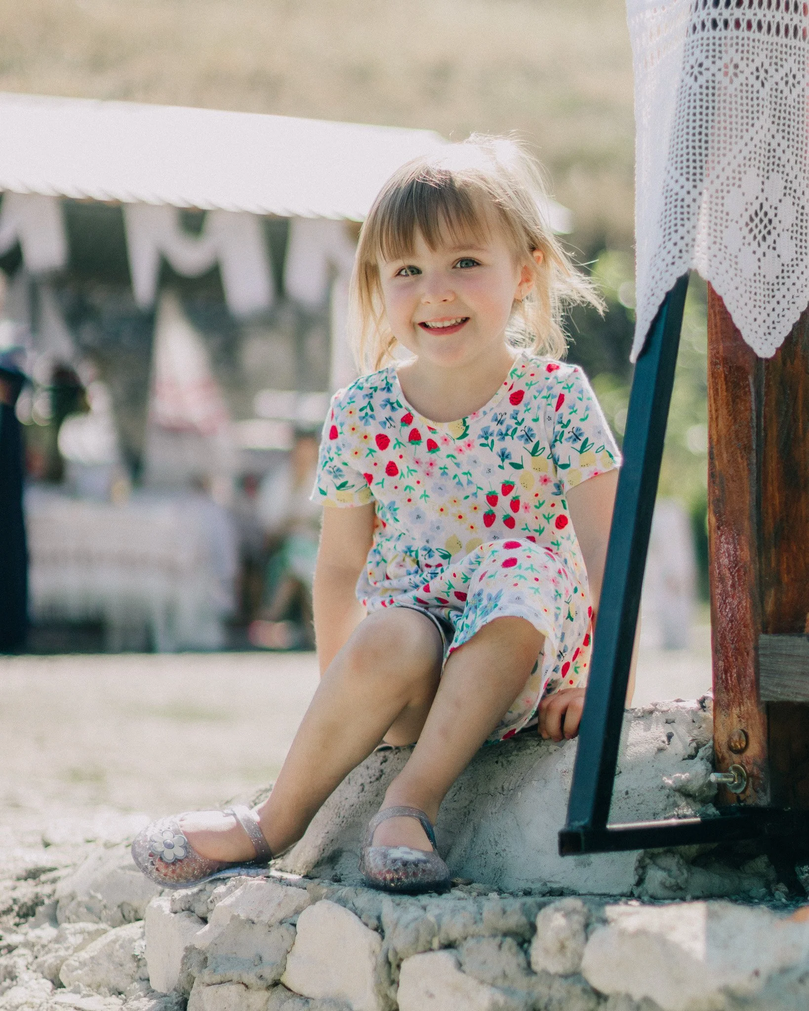A young girl with blonde hair and a colorful floral dress sitting cross-legged on a stone surface outdoors, smiling at the camera. In the background, there are blurred outdoor decorations and a banner.