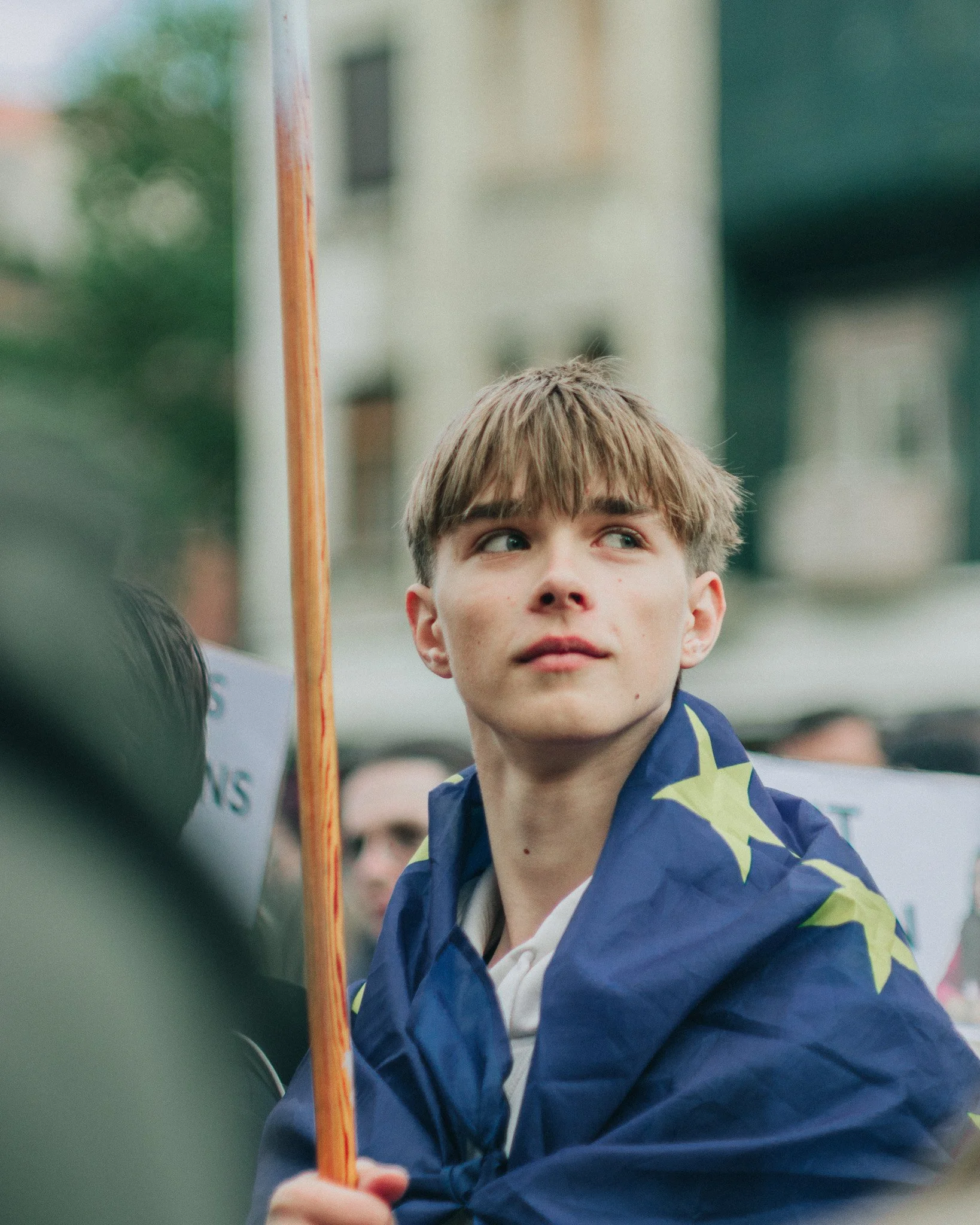 Young man with brown hair and light skin holding a flag with a blue background and yellow stars, possibly representing the European Union, participating in a public demonstration or march.