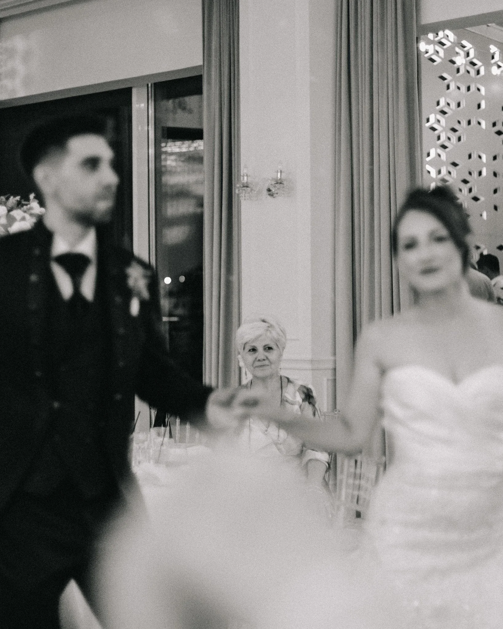 An elderly woman seated at a formal event, looking at a bride and groom dancing in the foreground, in a black and white photograph.