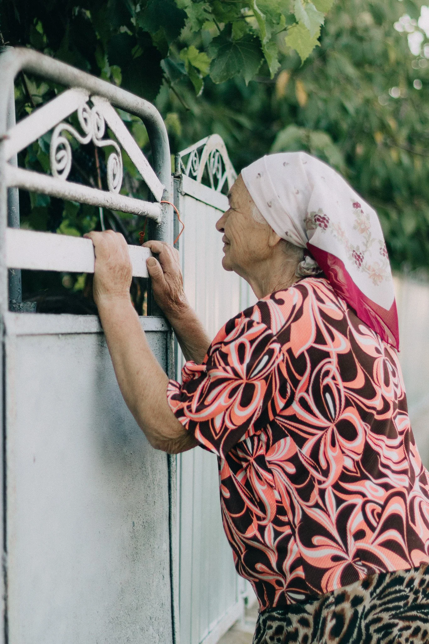 An elderly woman wearing a colorful patterned blouse and a floral headscarf, standing by a gate and smiling.