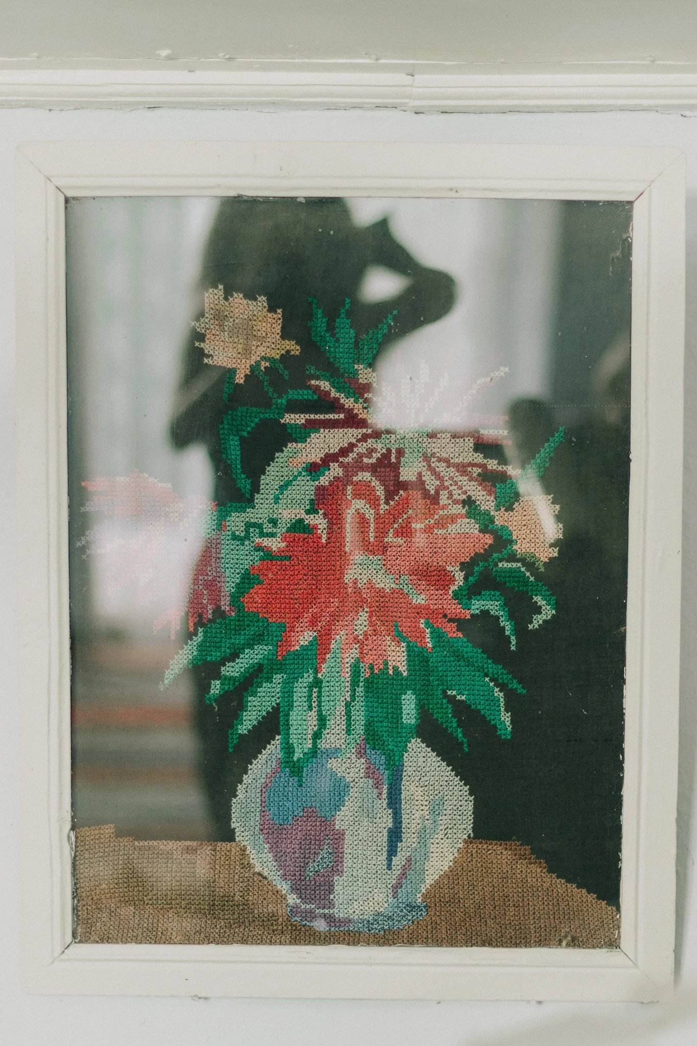 Cross-stitch embroidery of a poinsettia flower in a vase, viewed through a glass window.