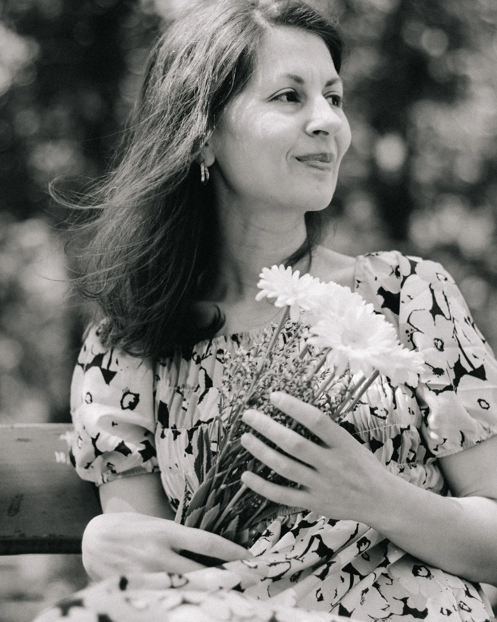 A woman with long hair wearing a patterned dress, holding a bouquet of flowers, sitting on a bench outdoors in a natural setting.