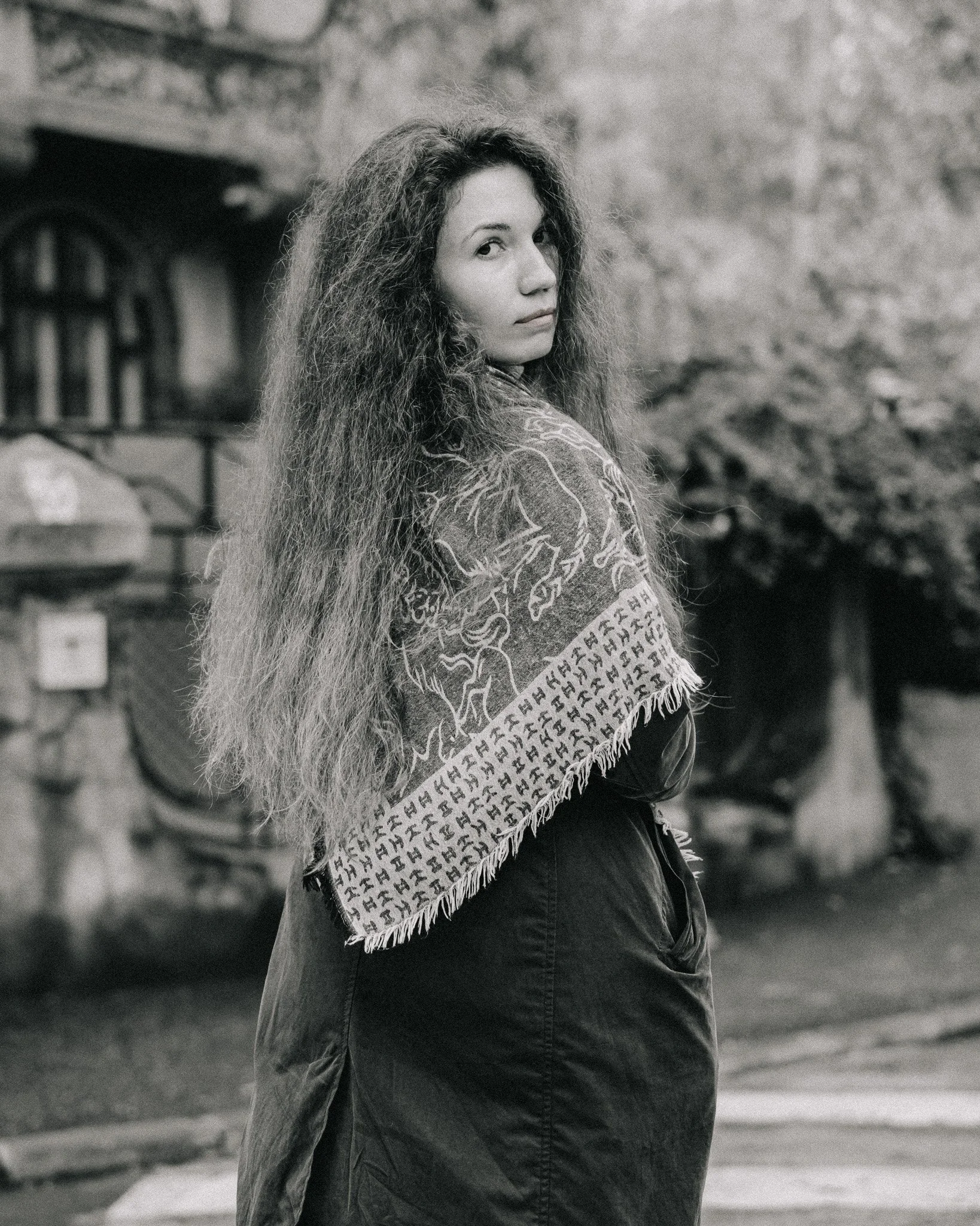 A woman with long, curly hair standing outdoors, looking over her shoulder towards the camera, wearing a patterned shawl and dark coat.