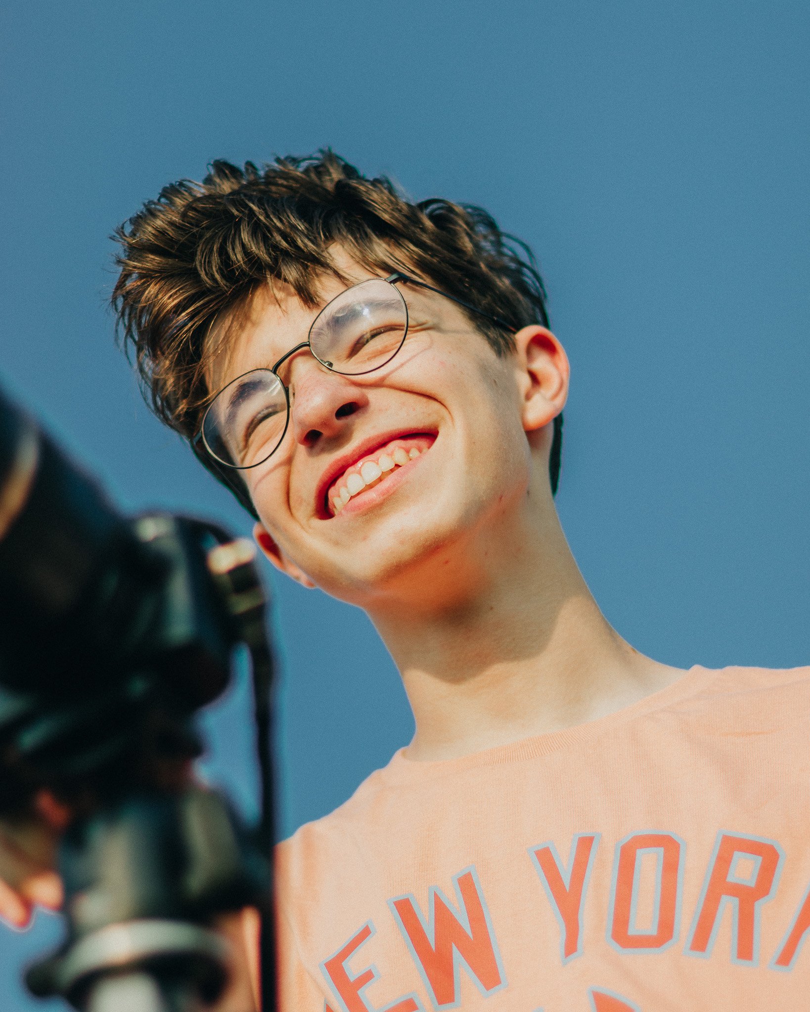Young man with glasses smiling and taking a photo outdoors under a clear blue sky