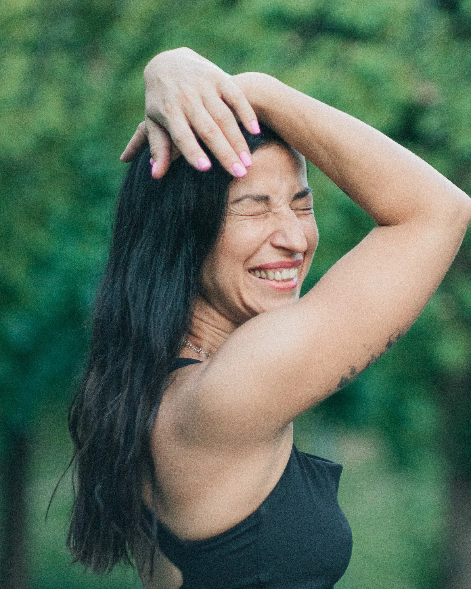 A woman with long black hair and a tattoo on her arm is smiling with her eyes closed and holding her head with one hand, outdoors with greenery in the background.