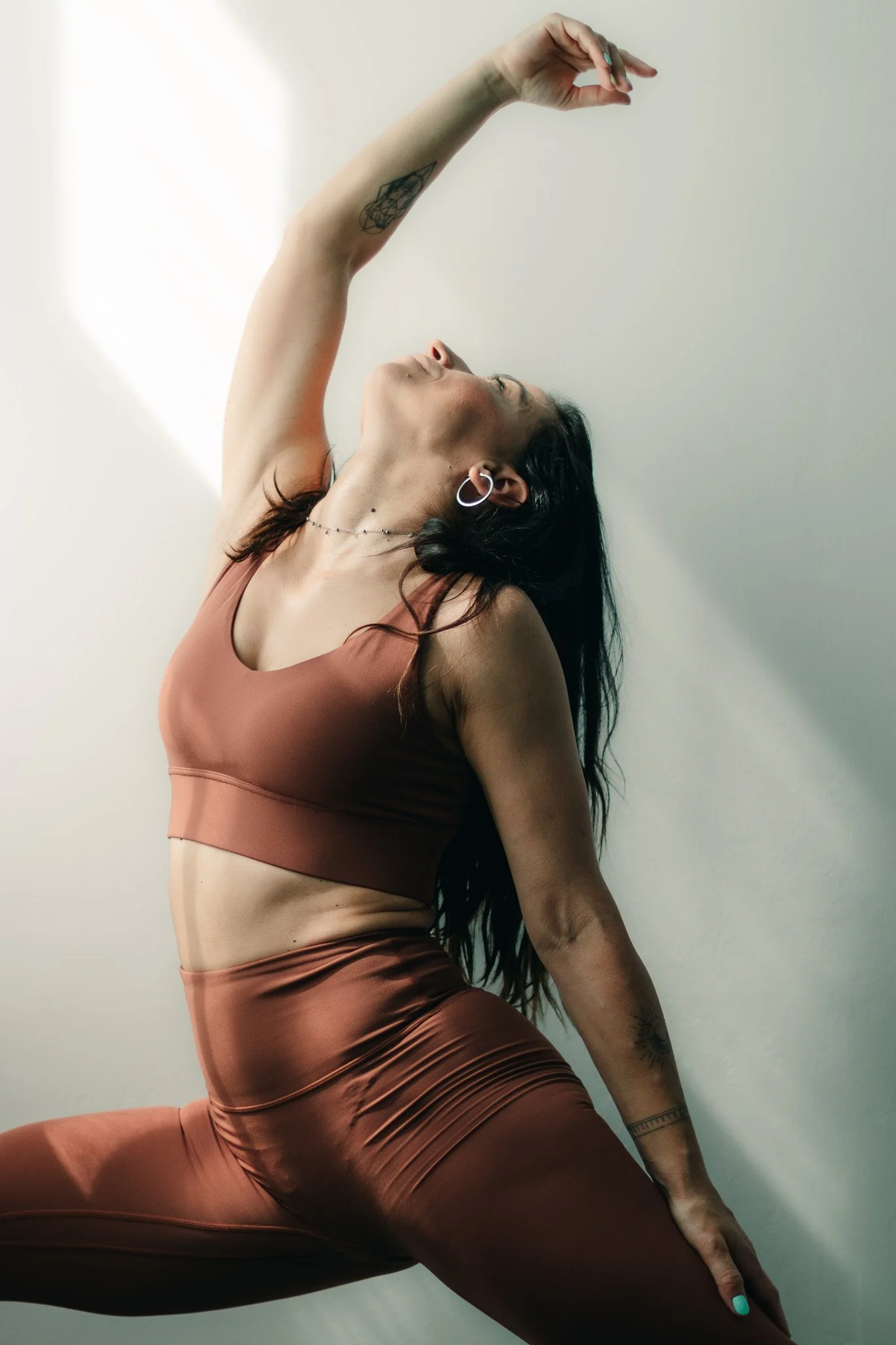 A woman with long dark hair practicing yoga against a plain wall, wearing a brown sports bra and matching high-waisted pants, with tattoos on her arm and wrist, in a side bend pose.
