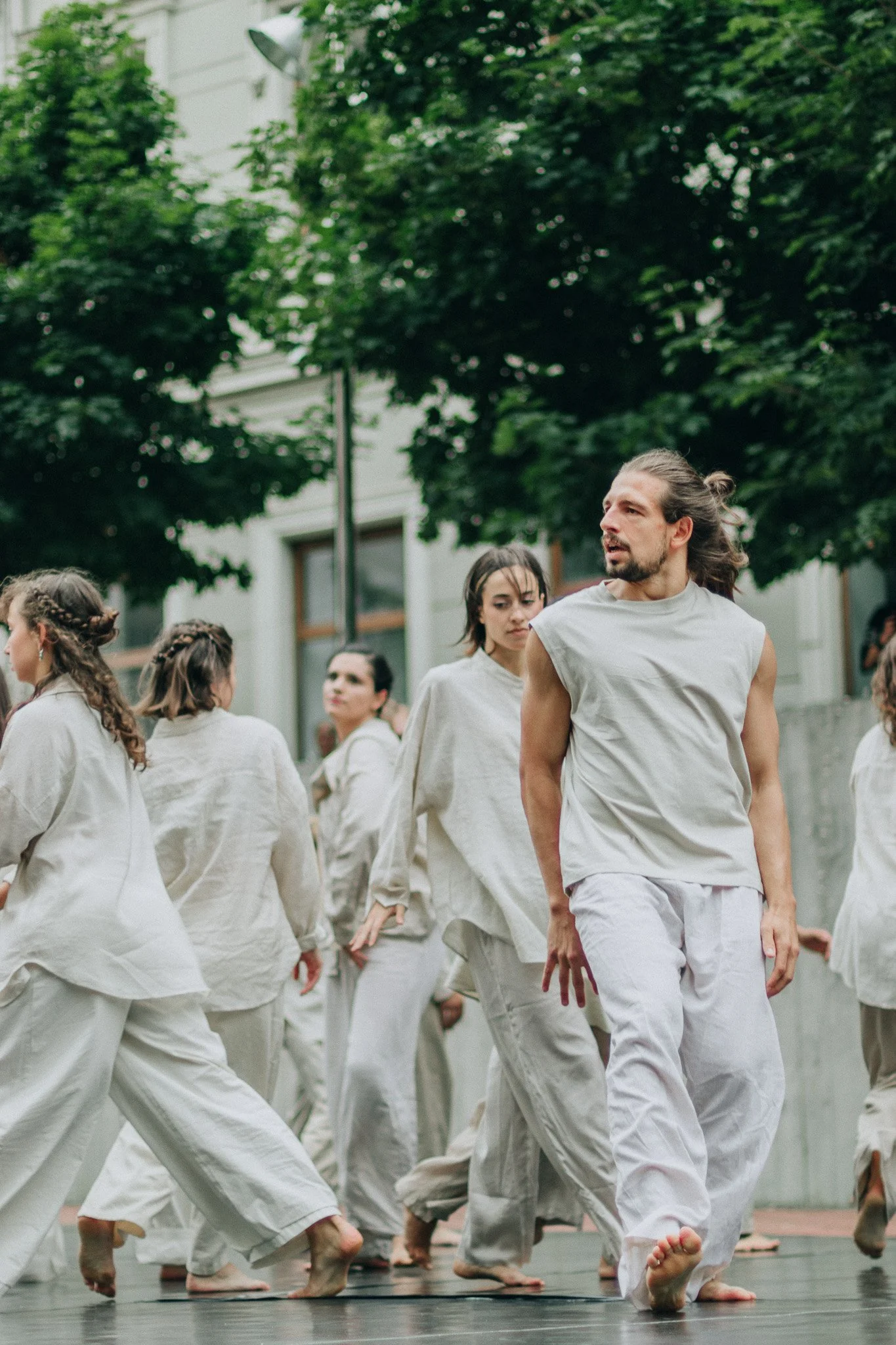 A group of people performing a dance outdoors on a wooden stage, dressed in white, with trees and a building in the background.