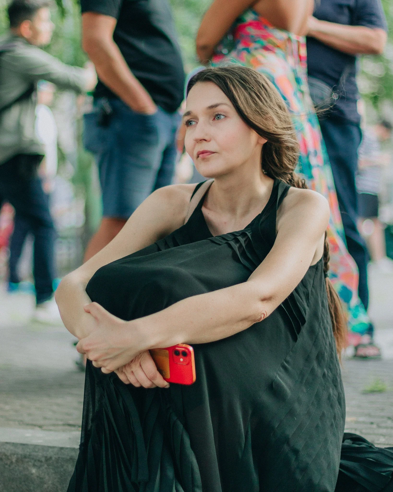 A woman sitting on the ground holding a black bag and a red phone, with people in the background standing and talking outdoors.