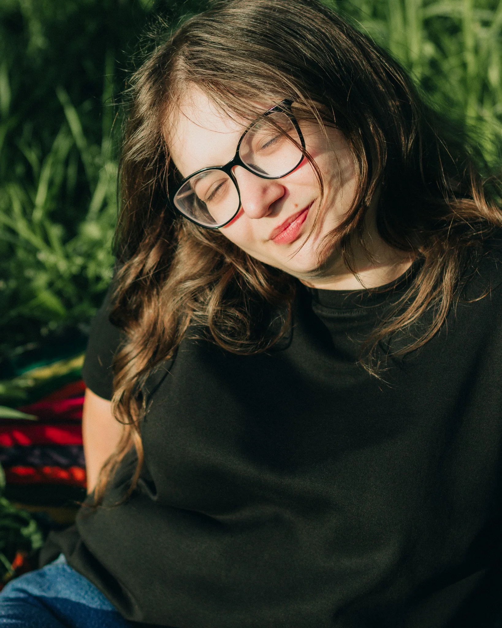 A woman with long brown hair and glasses sitting outdoors with her eyes closed and smiling, surrounded by green plants.