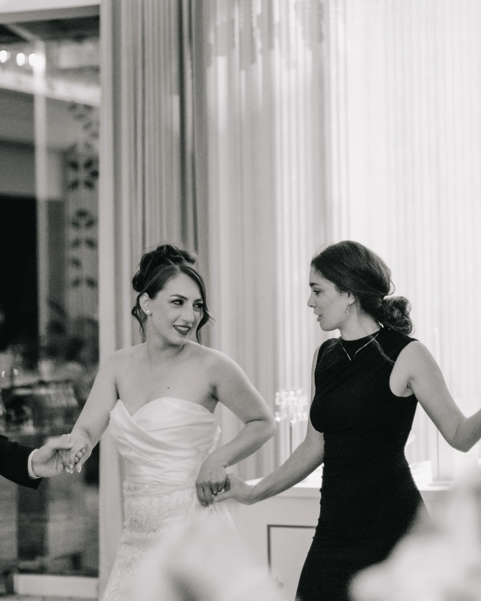 Two women holding hands and dancing at a formal event, one in a white dress and the other in a black dress, smiling and looking at each other.