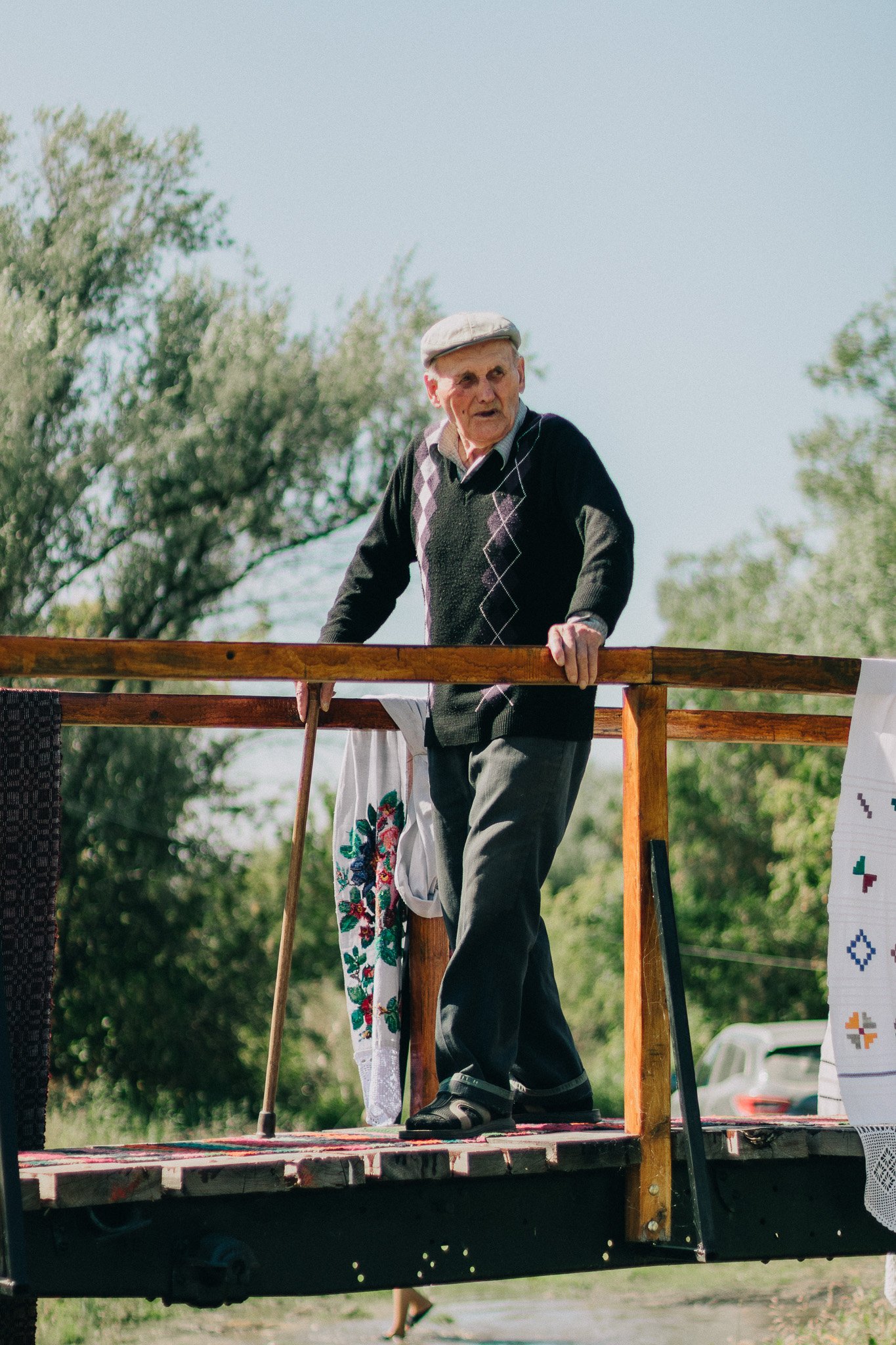 An elderly man standing on a wooden stage outdoors with greenery in the background, wearing a flat cap, a black sweater with a diamond pattern, and gray pants, holding a cane and looking to his left.
