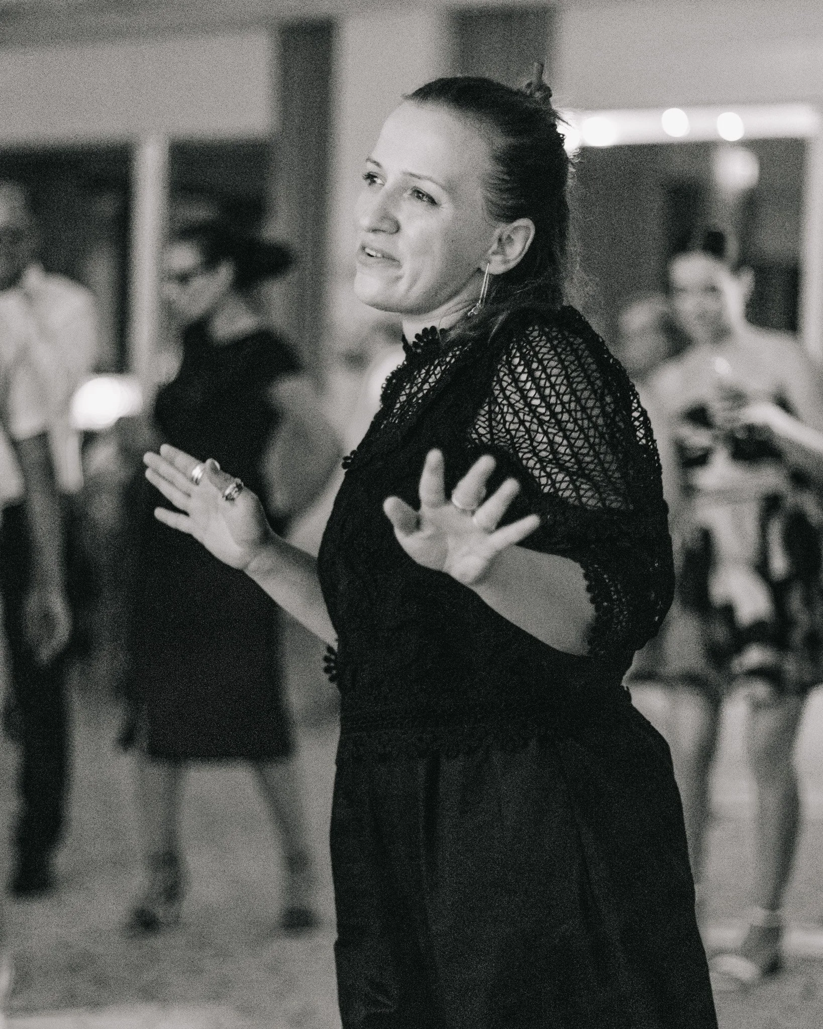 A woman in a black crocheted top dances with her hands raised and a smile at a social gathering, with people blurred in the background.