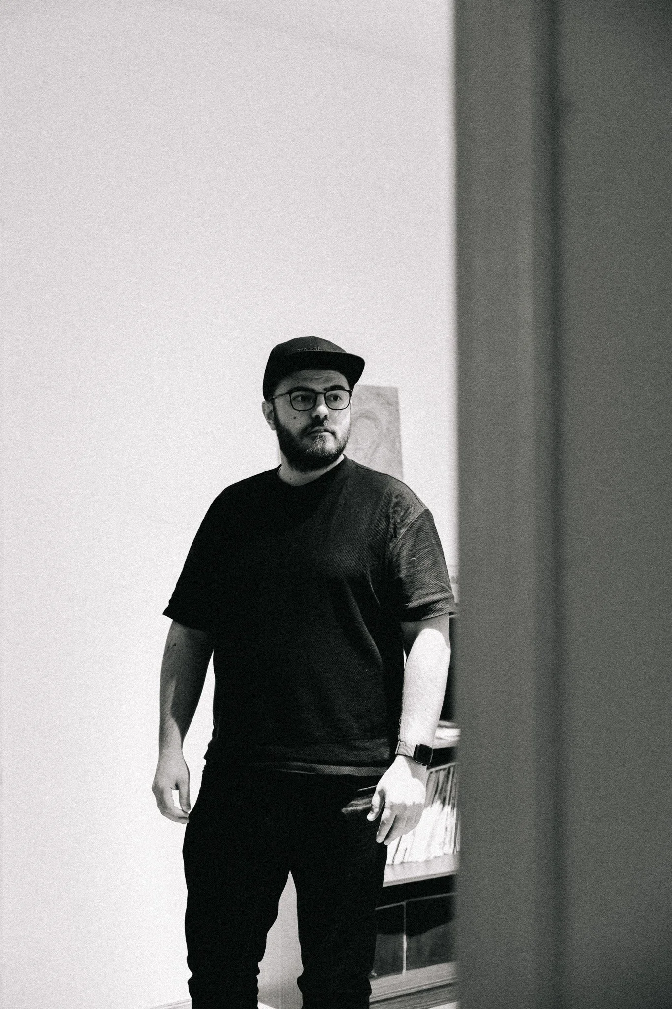 A black-and-white photo of a man with a beard, glasses, black hat, and black t-shirt standing indoors near a wall and a bookshelf.