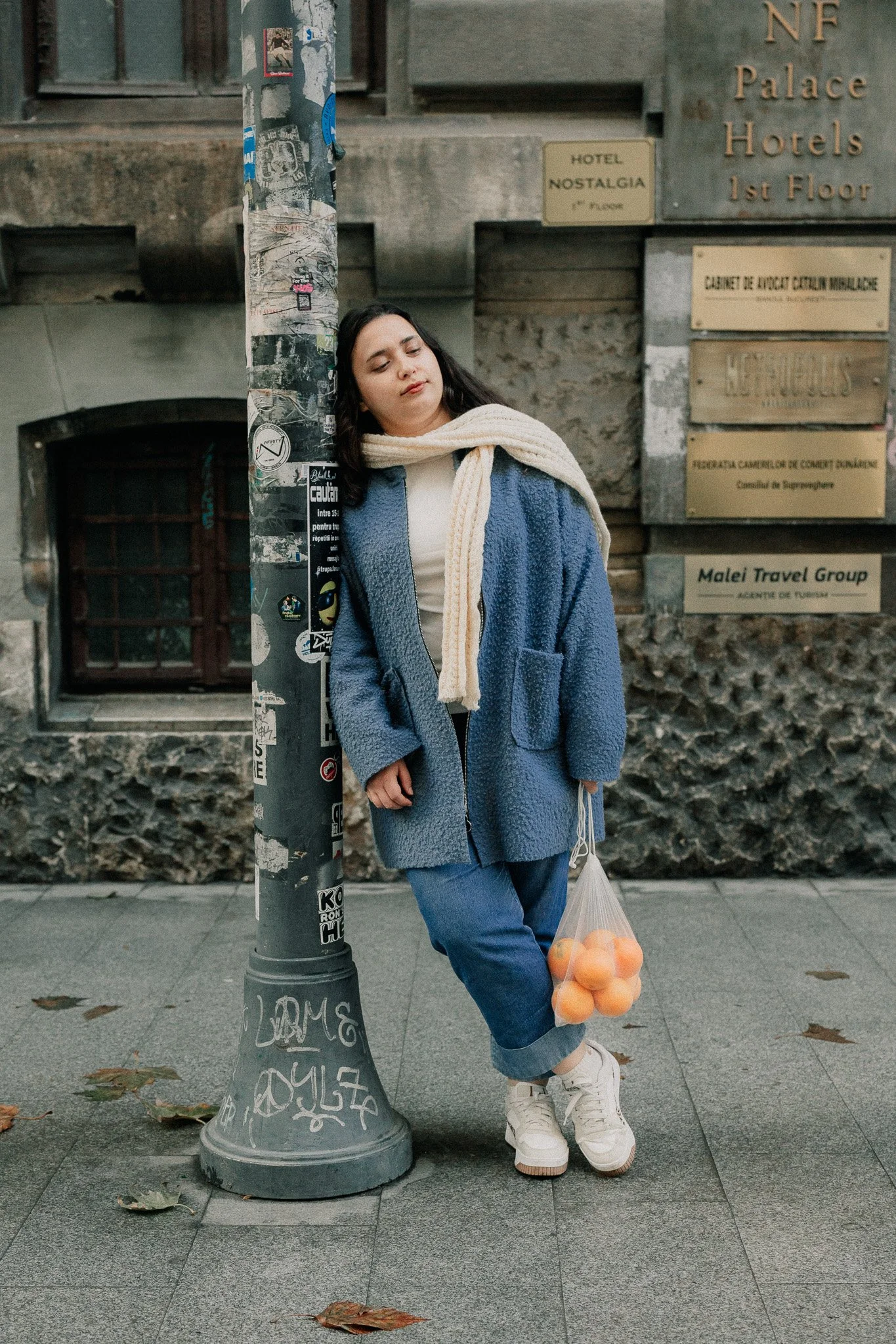 A young woman wearing a blue coat, white scarf, and white sneakers leaning against a pole with sticker decals on it. She is holding a plastic bag of oranges in her left hand, standing on a city sidewalk with fallen leaves. In the background, there is