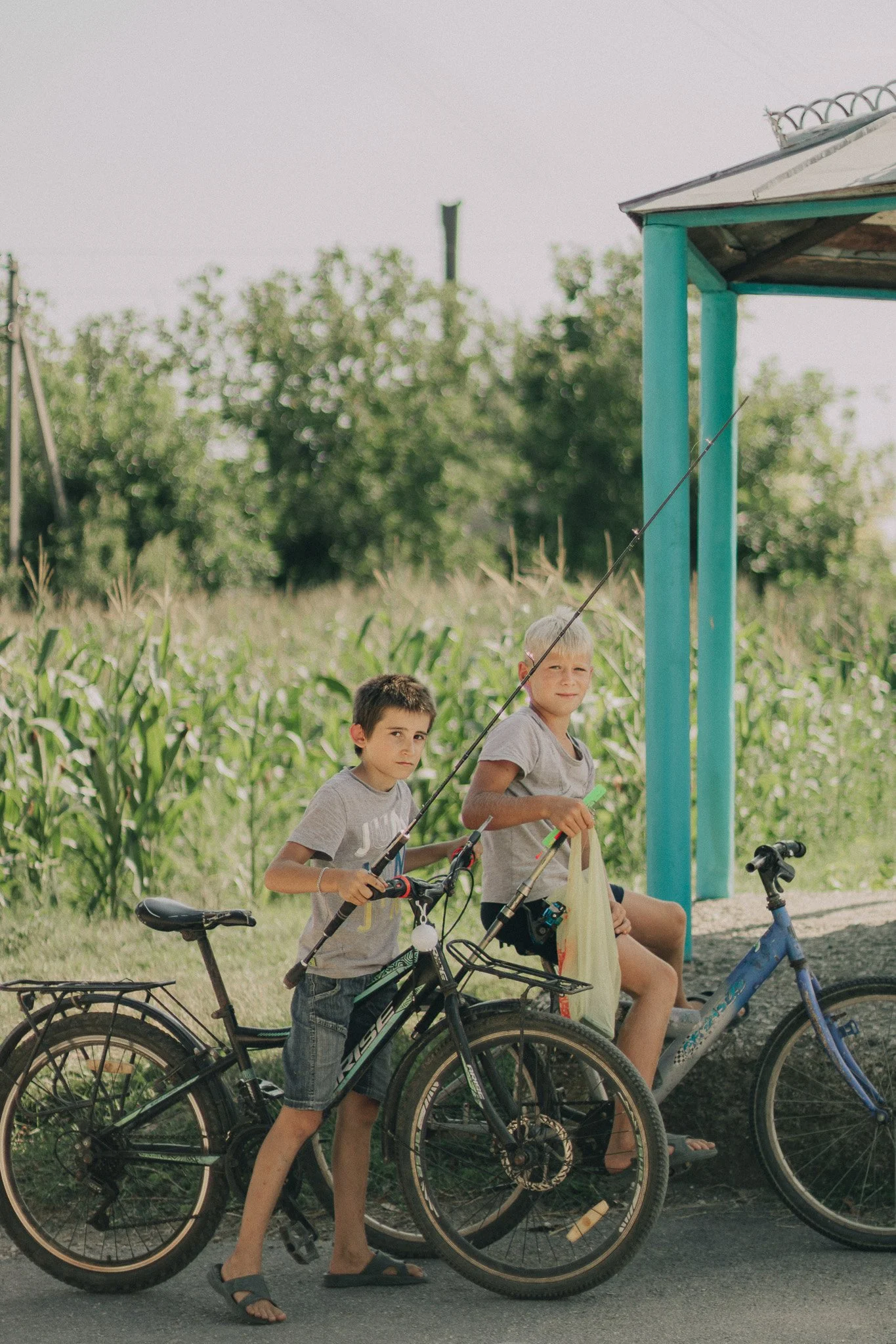 Two boys with bicycles near a small shelter in a rural area, with greenery and trees in the background.