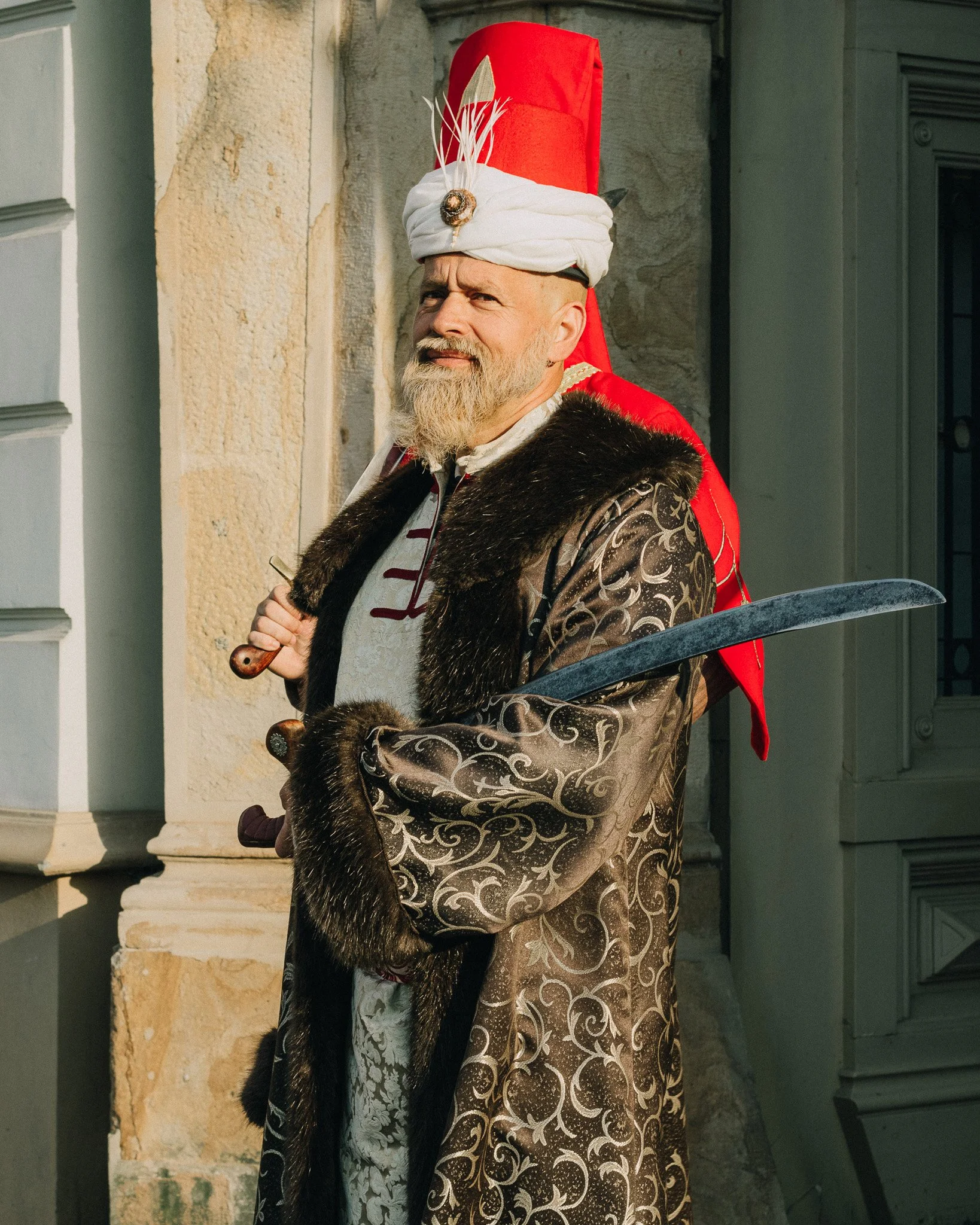 Man dressed in medieval-style clothing holding a sword, wearing a tall red hat with white fabric and feathers, standing outside a building.