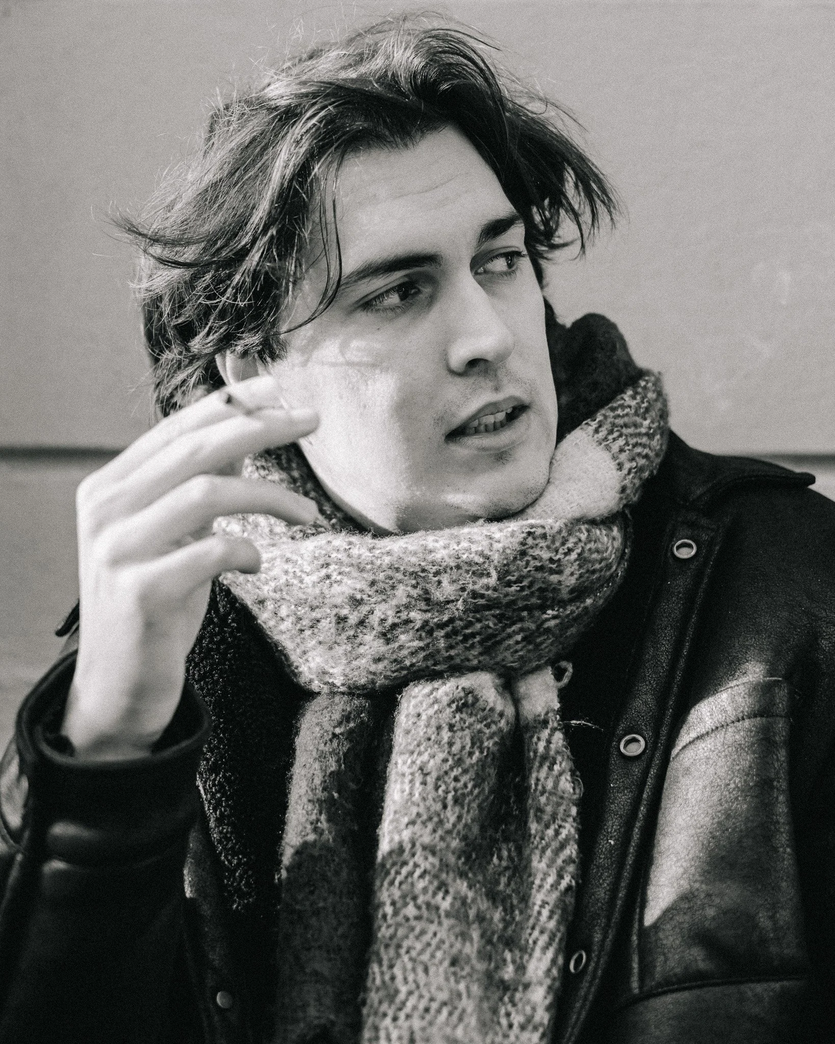 A young man with messy hair wearing a leather jacket and a thick scarf, looking to his left with a contemplative expression.
