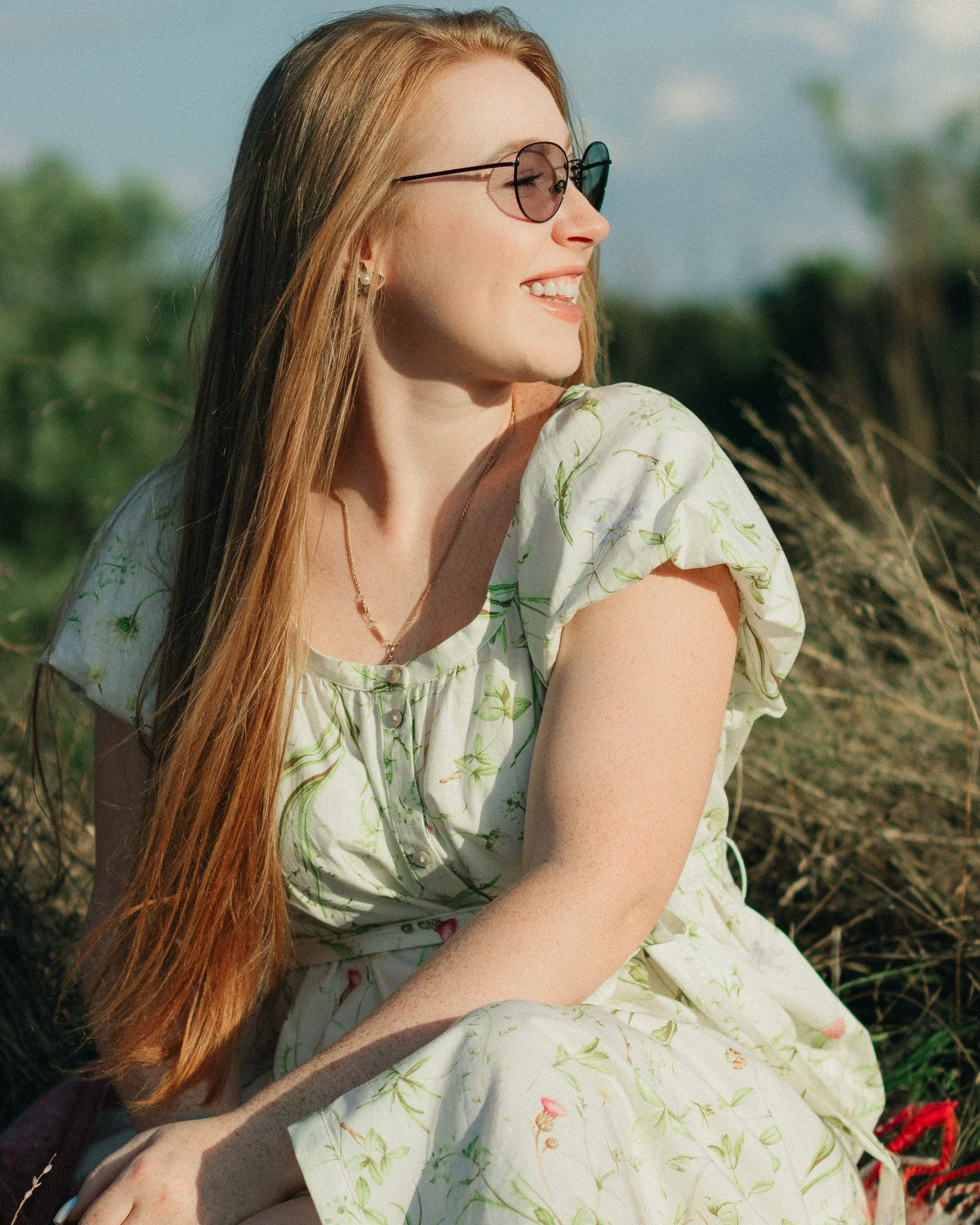 Young woman with long red hair, sunglasses, and a floral dress, sitting outdoors in a field with tall grass, smiling and looking to the side.