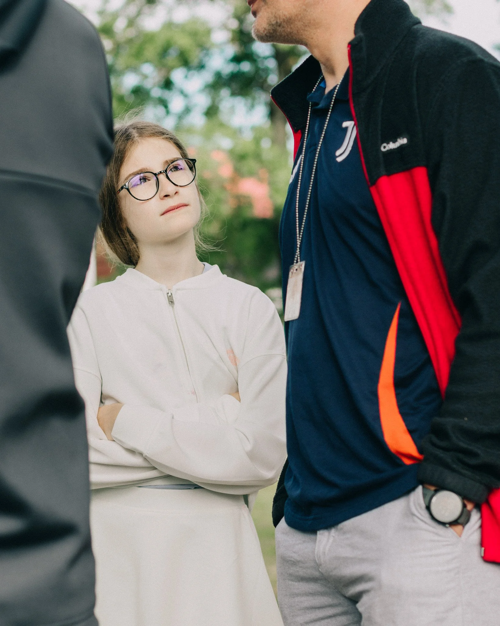 A young girl with glasses and long brown hair listening intently to a man while standing outdoors. The girl is wearing a white jacket, and the man is wearing a navy blue and red jacket with a lanyard around his neck. Another person is partially visib