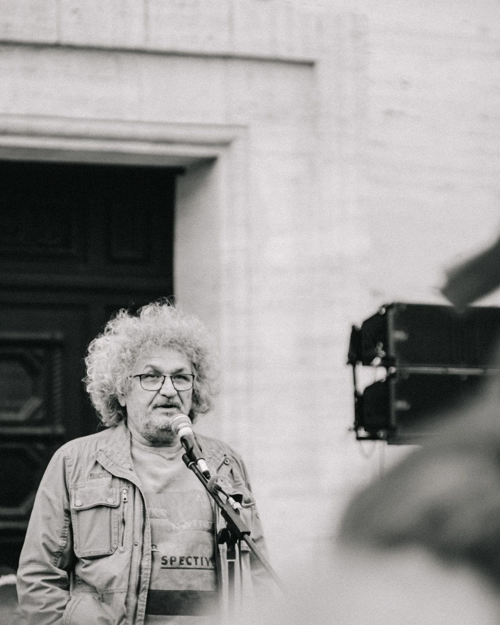 A man with curly white hair and glasses speaking into a microphone during a presentation, in front of a large speaker and a brick wall.