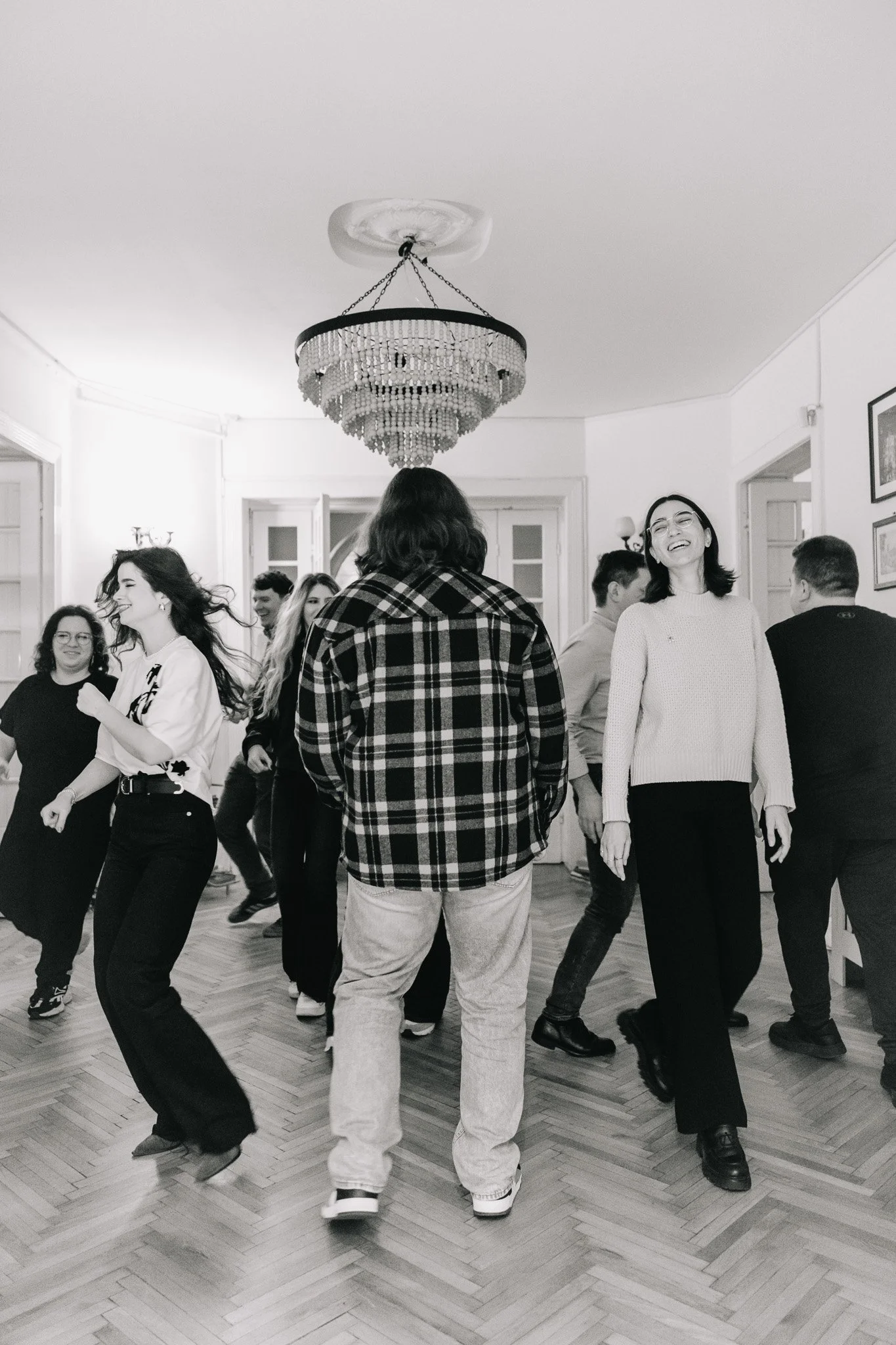 People dancing and laughing in a living room with wooden floors and a chandelier.