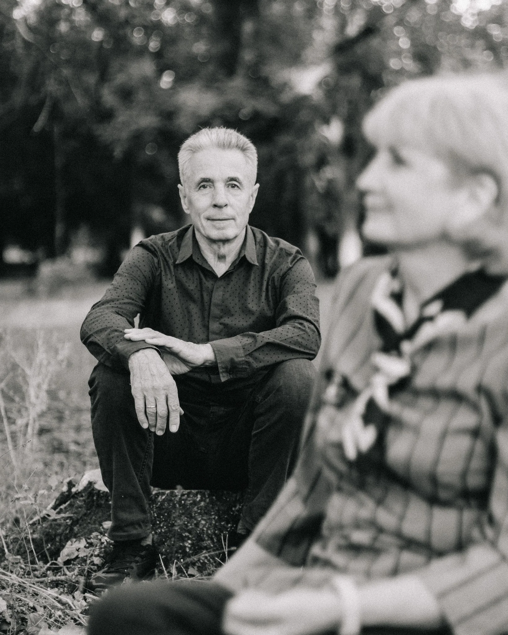 Black and white photo of a man sitting on the ground outdoors, with trees in the background. He is looking directly at the camera with his arm resting on his knee. A woman with glasses and short hair, wearing a plaid shirt, is sitting nearby and is o