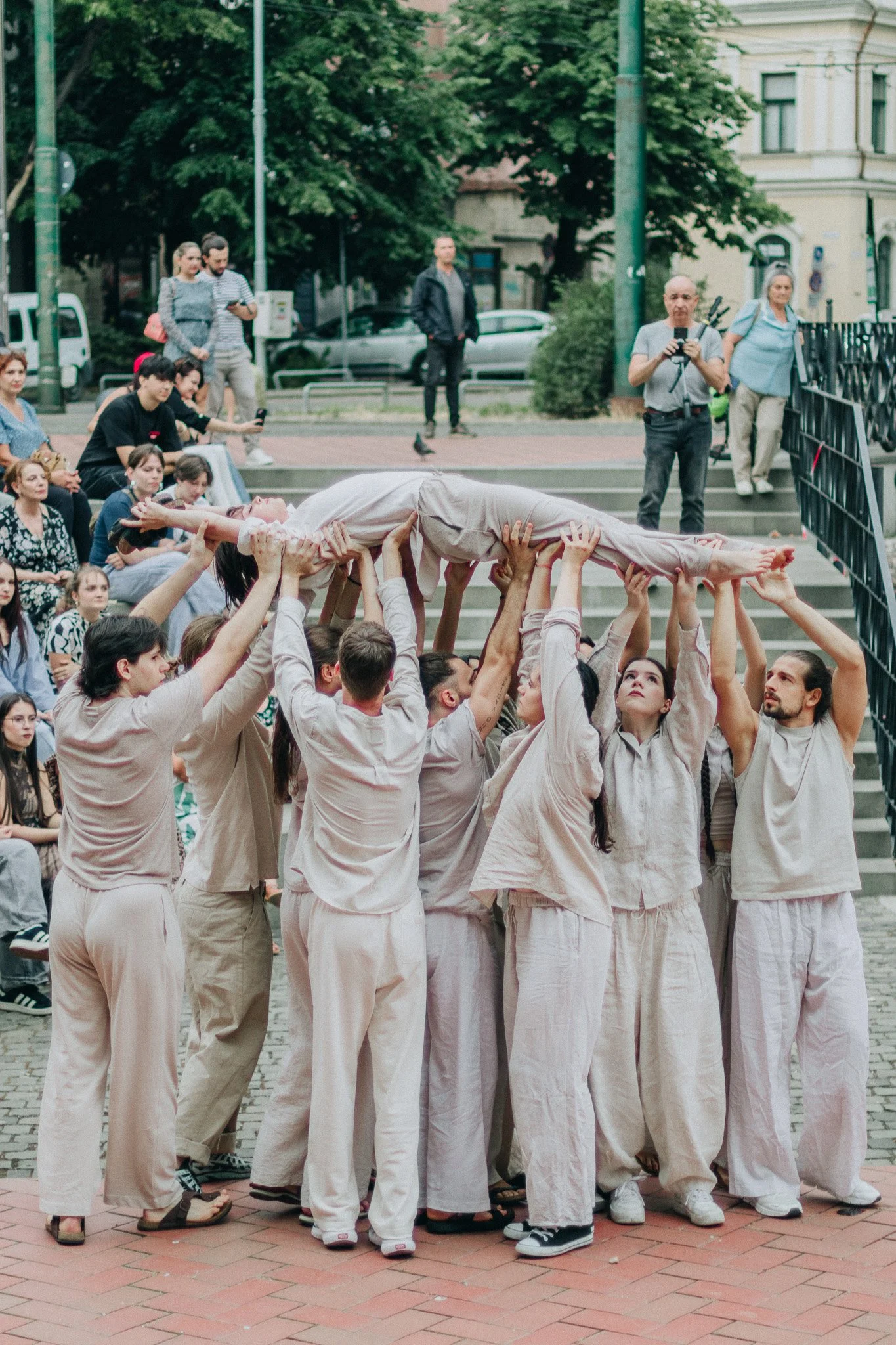A group of people dressed in beige clothing perform a coordinated human tower outdoors in front of an audience seated on steps and standing nearby. Some audience members are taking photos or videos.