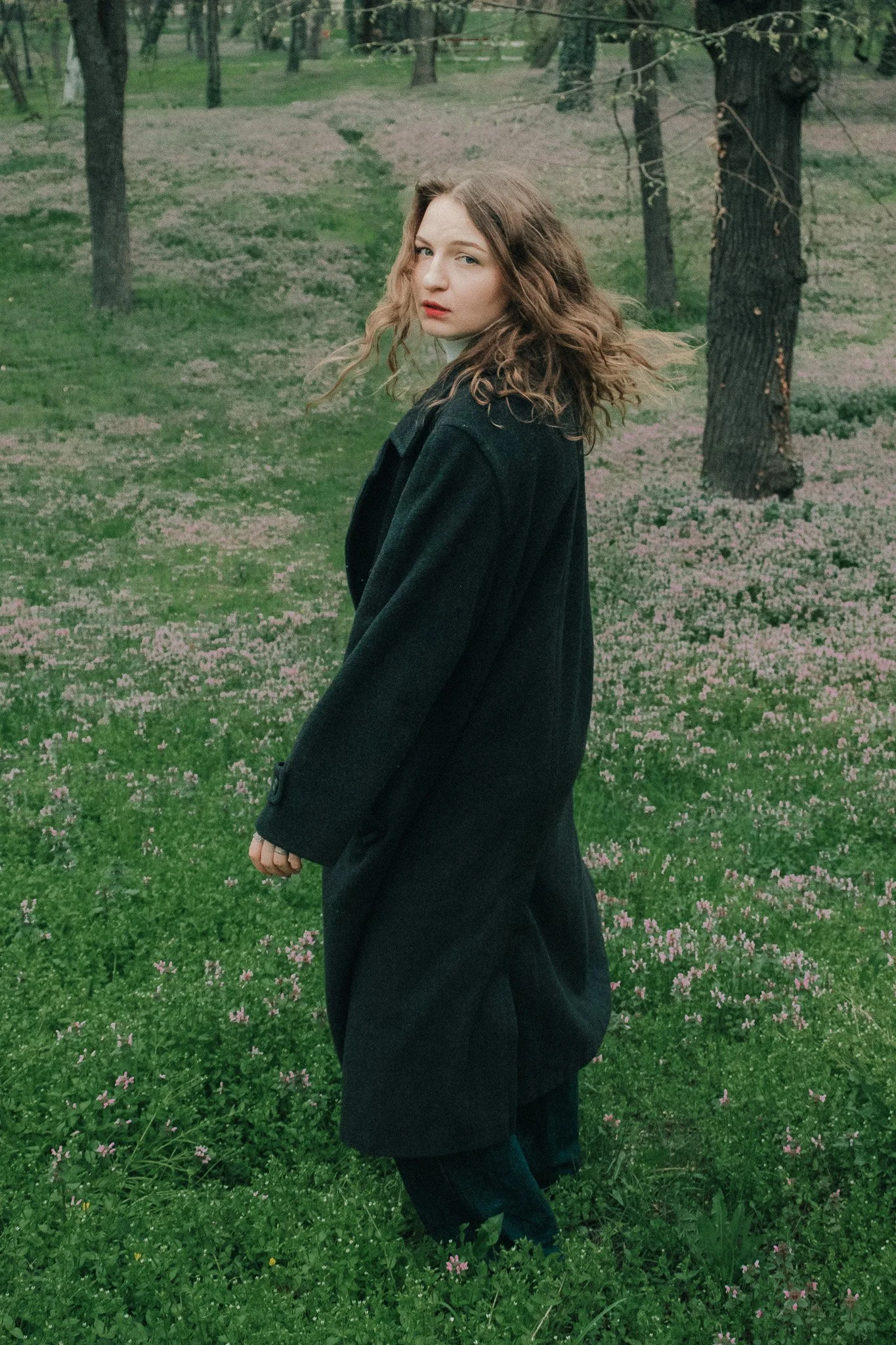 A young woman with curly brown hair stands in a park with pink flowers and trees, wearing a black coat.
